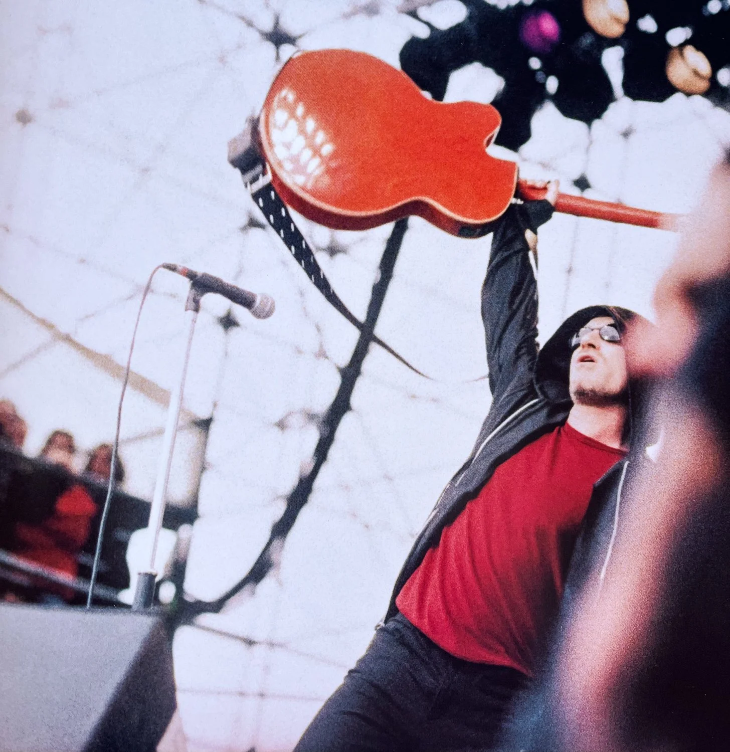 A person with long dark hair, wearing sunglasses, a red shirt, and a black jacket, holding an orange ukulele up in the air during a live performance, with a microphone and a group of people visible in the background.