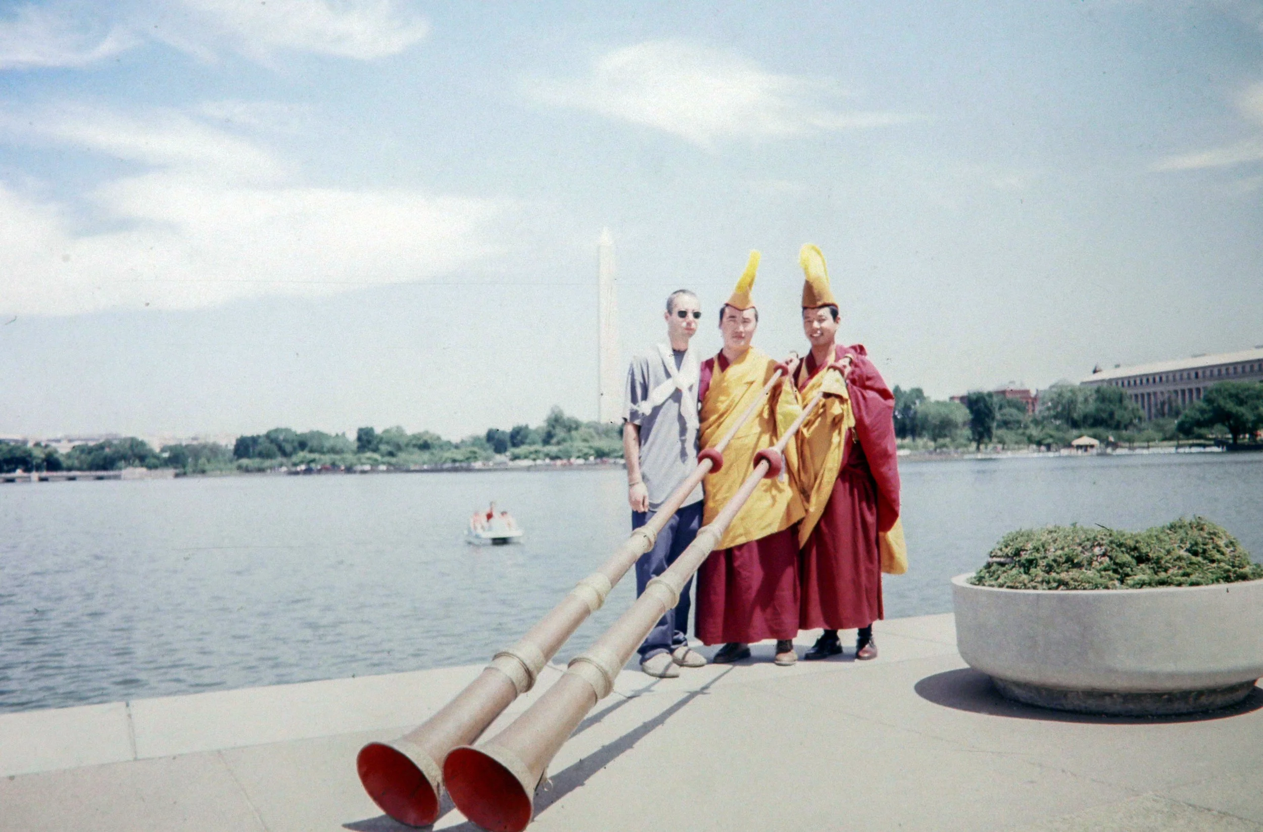 Three individuals standing by a body of water, with two of them dressed as traditional monks in maroon and yellow robes, wearing large yellow hats, and playing long musical horns. A third person in casual clothing is standing beside them. In the background, there is a boat on the water, trees, and buildings.