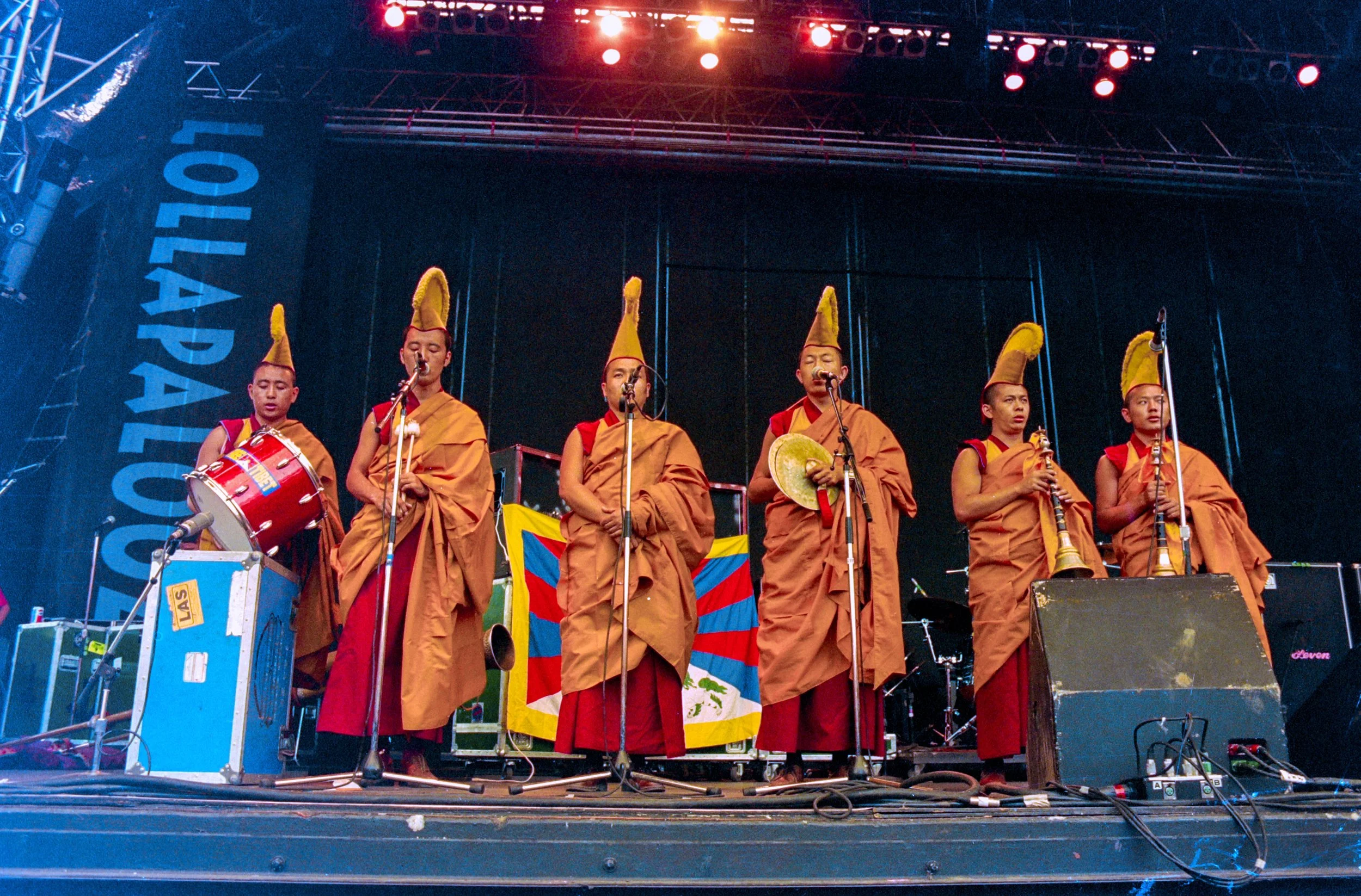Group of monks dressed in traditional robes and hats performing at a stage, with musical instruments and microphones, during an outdoor cultural event.