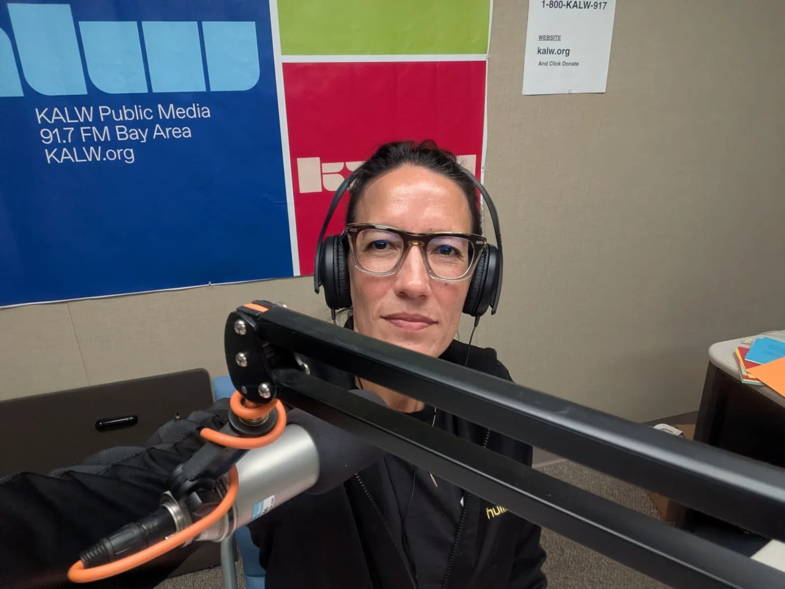 A woman wearing glasses and headphones sits in a recording studio with KALW public media branding behind her.