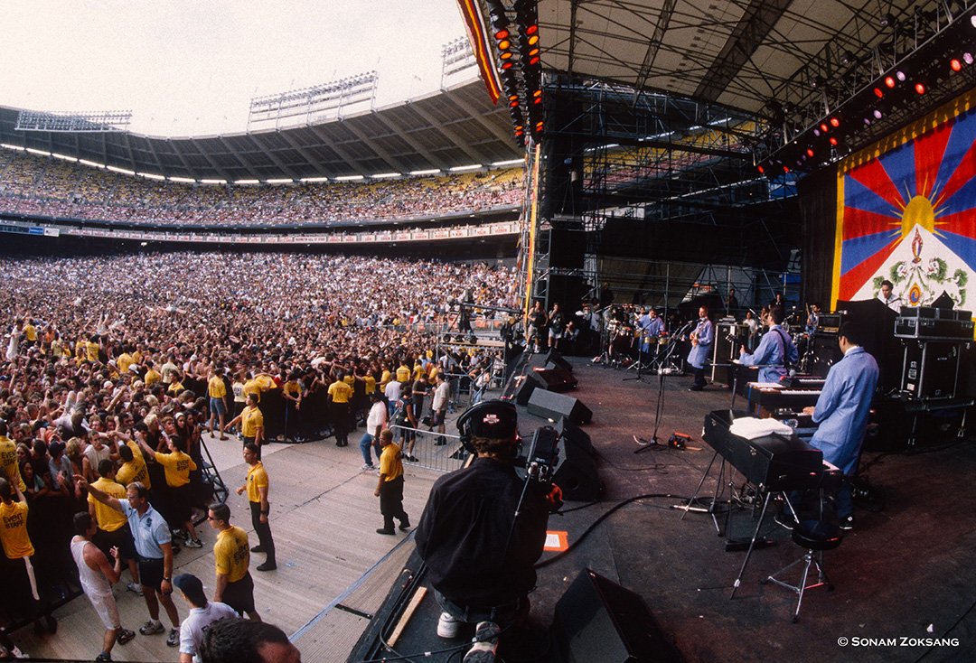 A wide view of the 1998 Tibetan Freedom Concert at RFK Stadium in Washington, D.C., showing the stage, performers, big screen, security staff, and a large crowd.