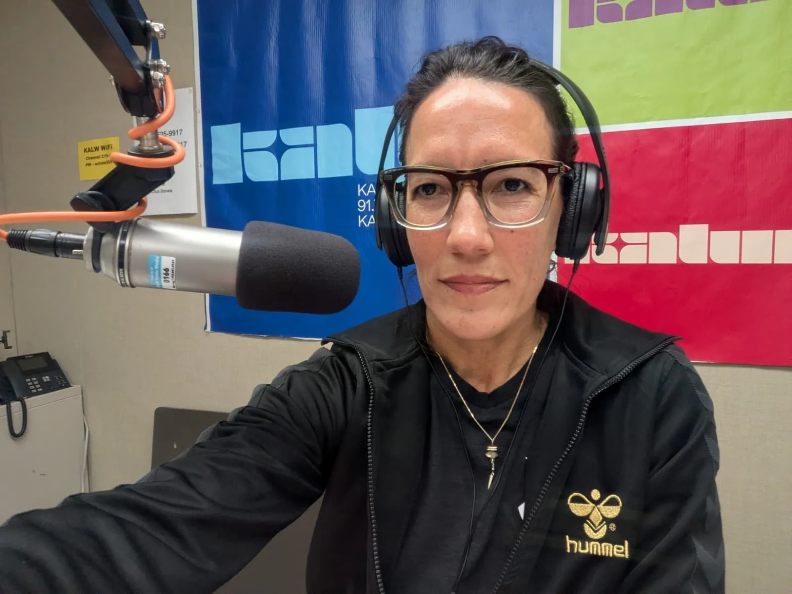 A woman in glasses is taking a selfie in a recording studio. She is wearing headphones and a black jacket with a logo, and there are colorful banners behind her.