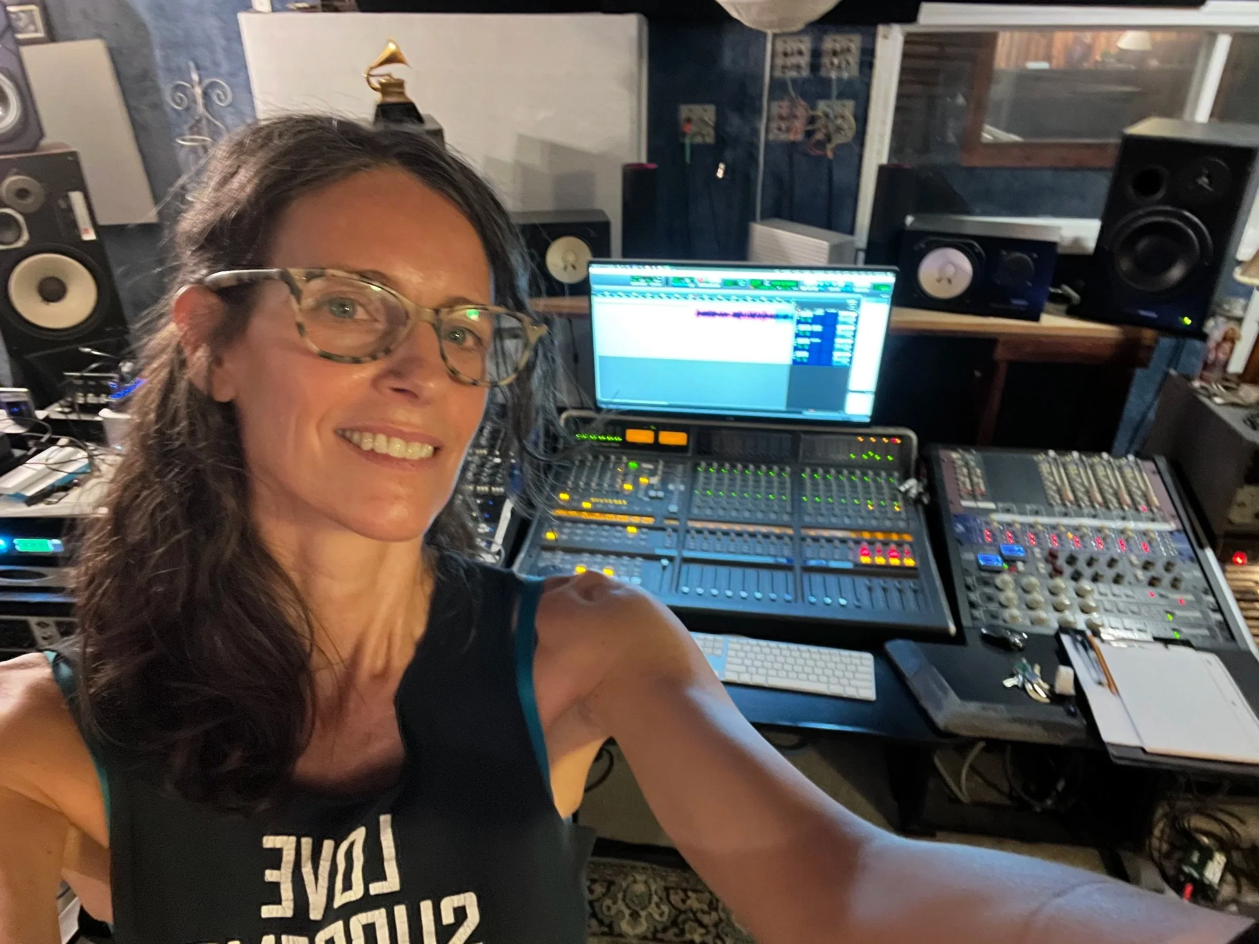 A woman with glasses smiling taking a selfie in a music studio with audio equipment, mixing desk, speakers, and a computer screen displaying music editing software.