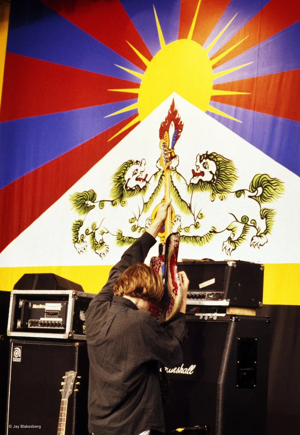A person with red hair playing an electric guitar in front of a colorful mural with a sun and lion motifs, with audio equipment nearby.