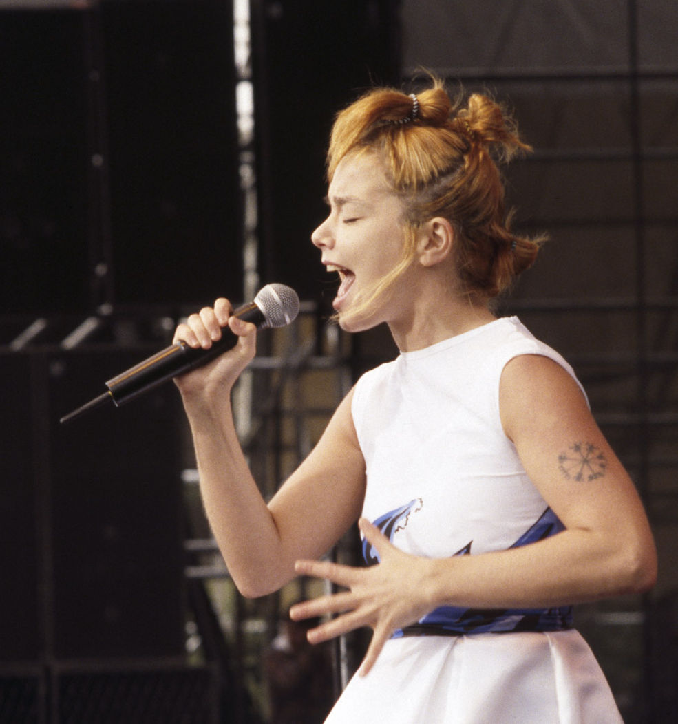 A woman with short, messy, reddish-blonde hair is singing into a microphone on stage, with one hand on her stomach and the other holding the microphone.