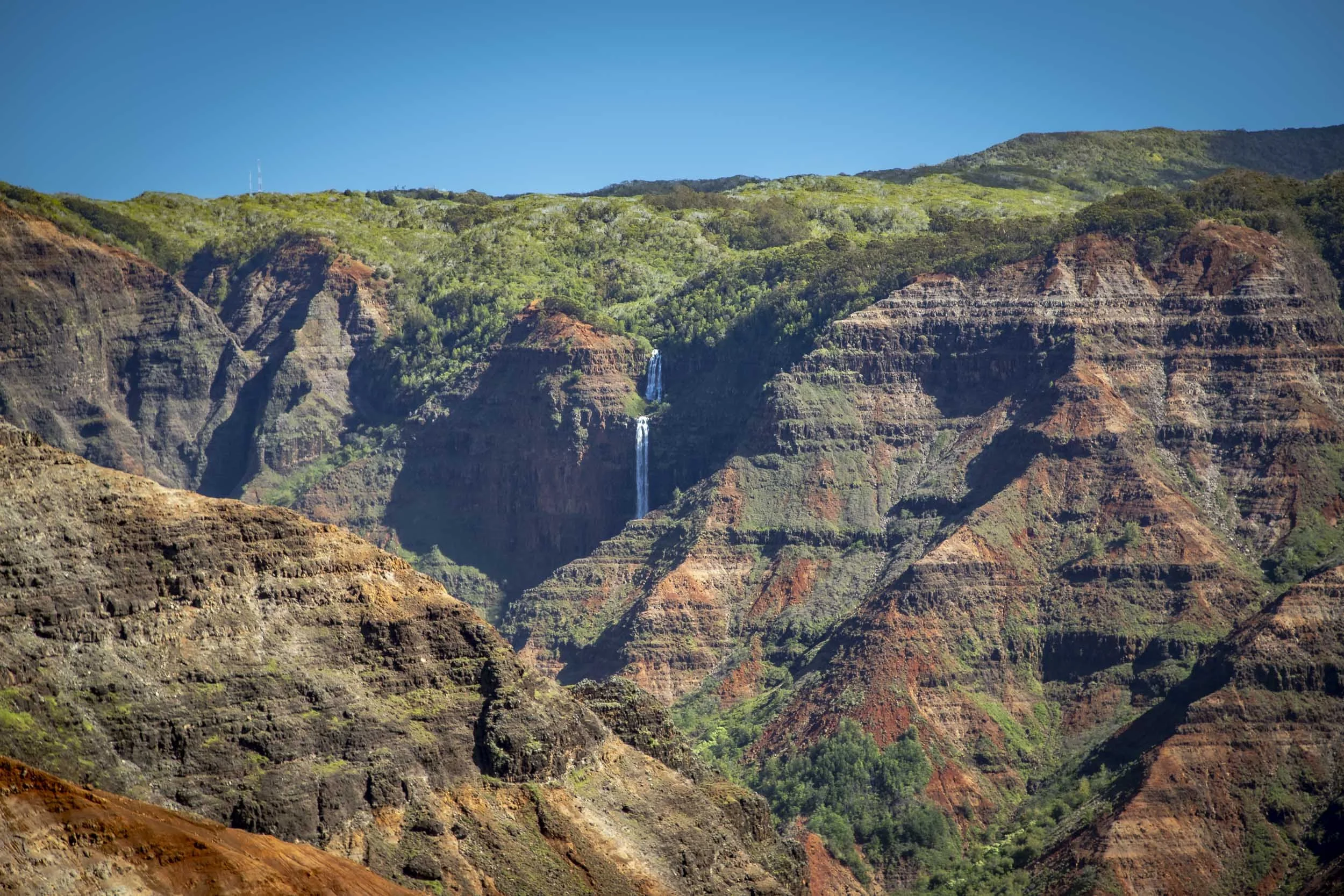 Waterfall cascading down the rugged red and green cliffs of a vast canyon under a clear blue sky. Location: Waimea Canyon, Hawaii.