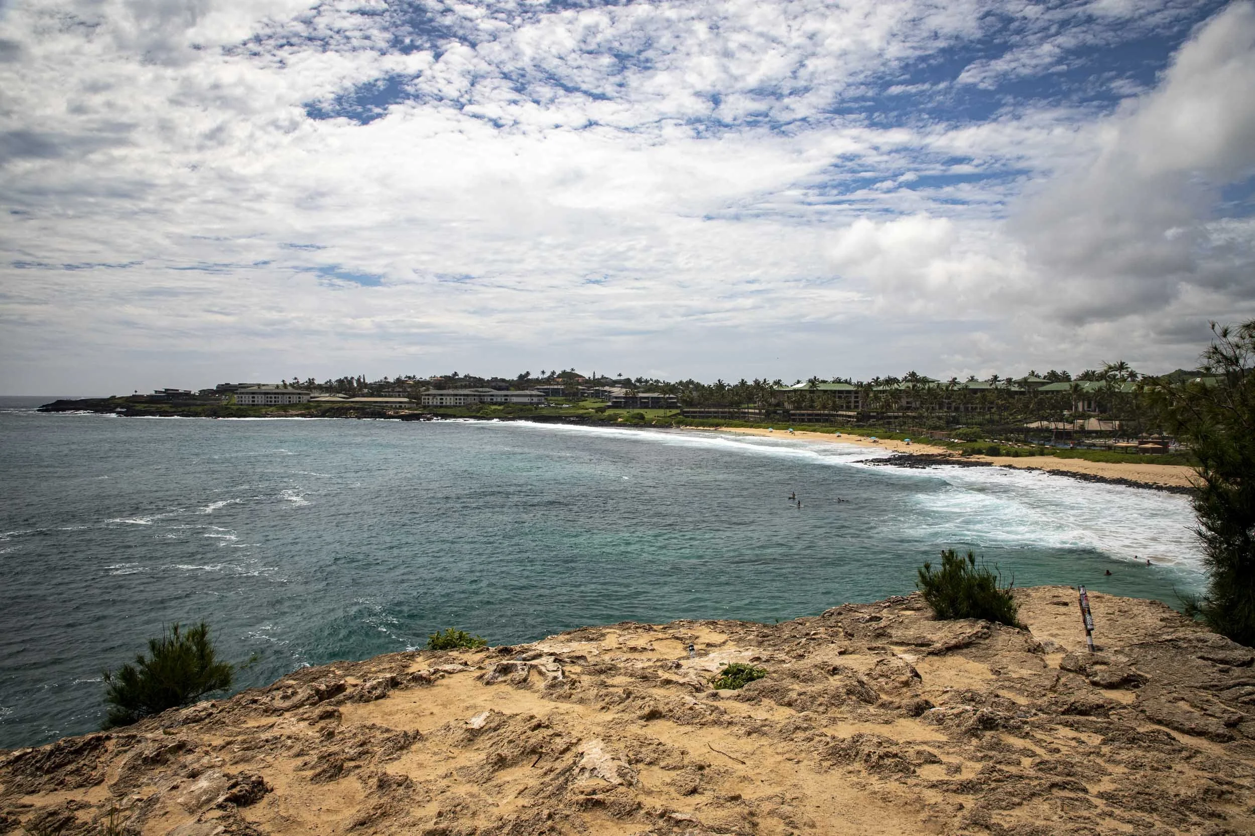 Rocky cliff overlooking a turquoise bay with waves and resorts along a sandy shore. Shipwreck Beach, Kauai, Hawaii.