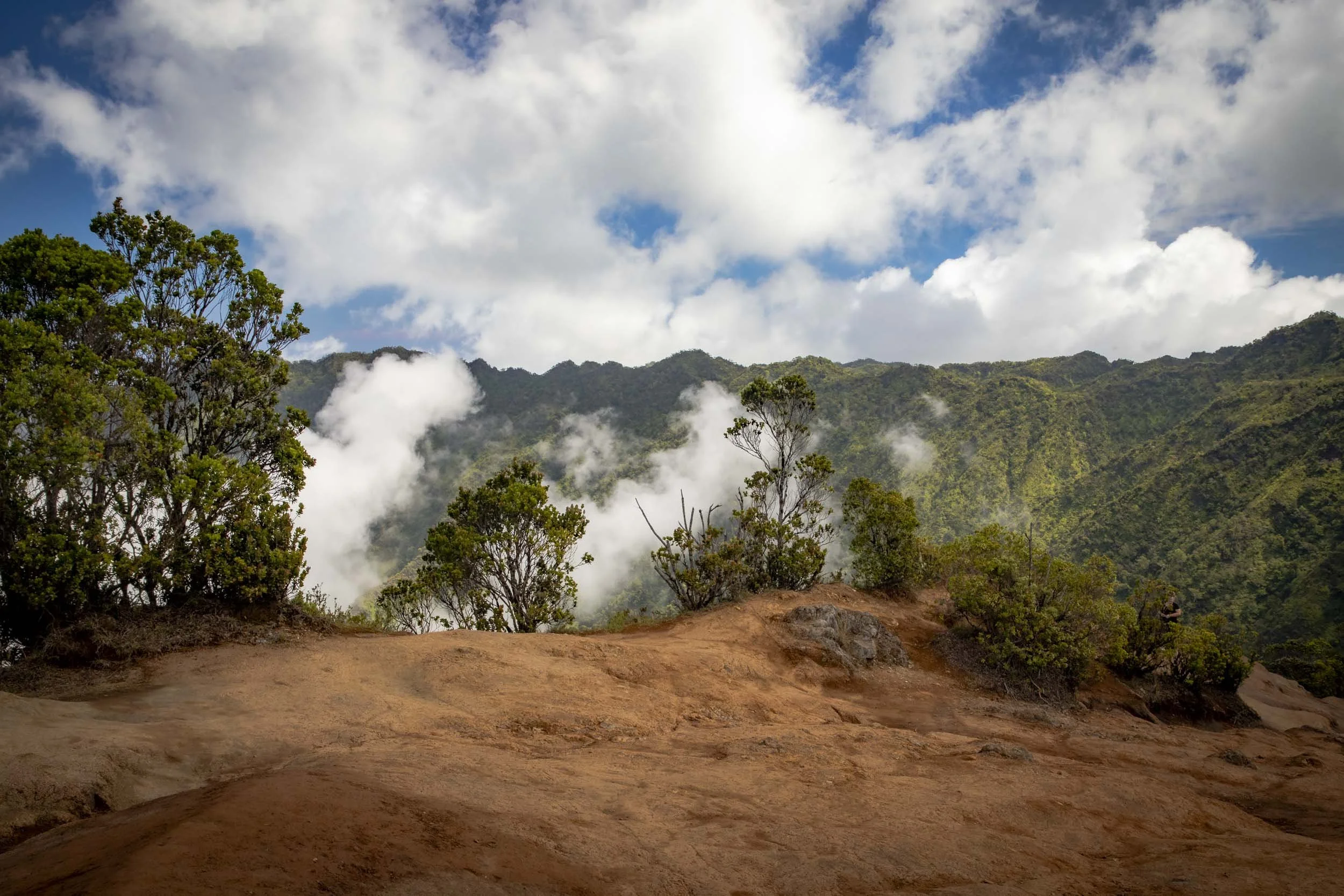 Red rocky overlook with scattered trees, clouds drifting over lush green mountains. Location: Waimea North Lookout, Hawaii.