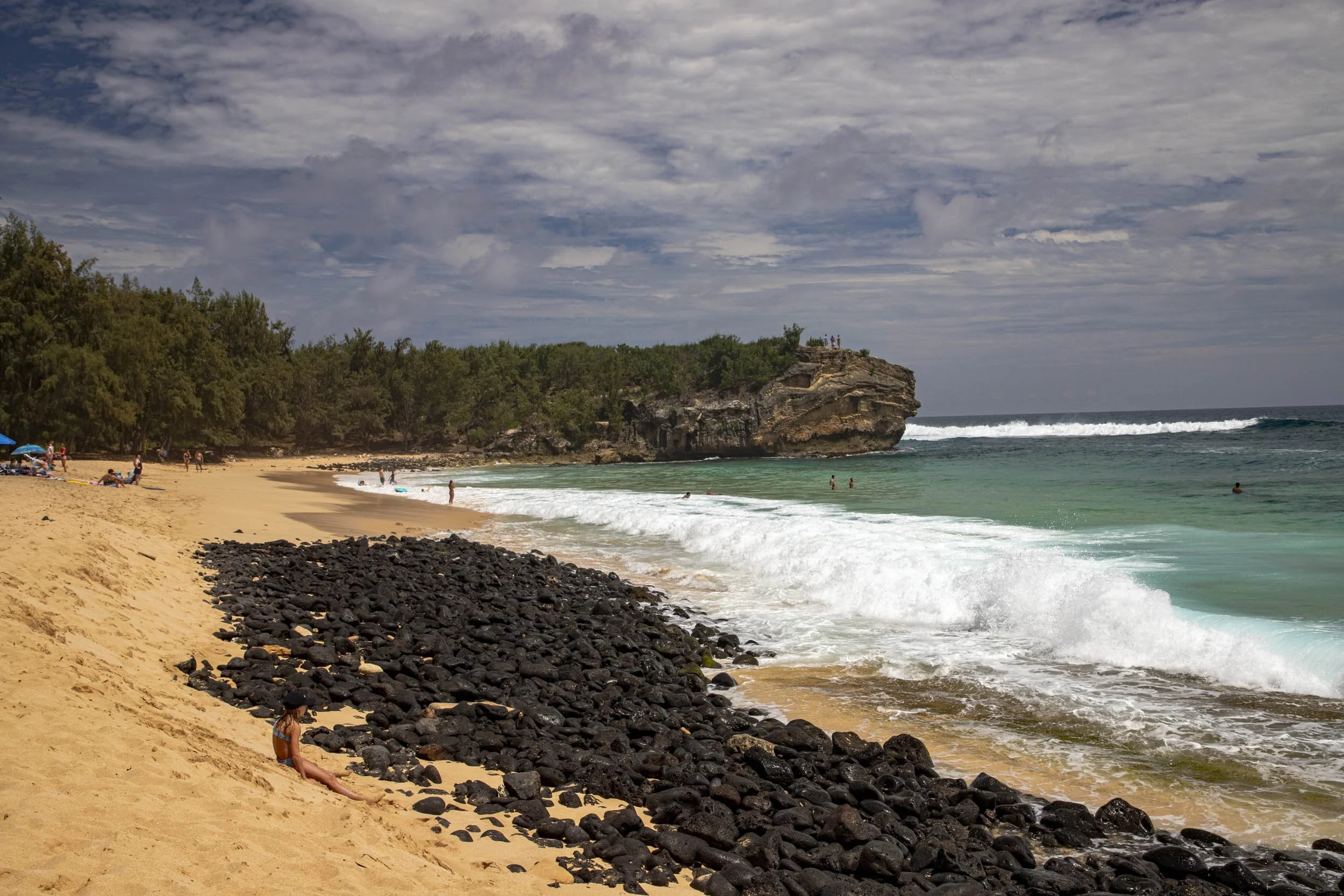 Golden sandy beach with dark lava rocks, turquoise waves, and a rocky cliff under a partly cloudy sky. Shipwreck Beach, Kauai.