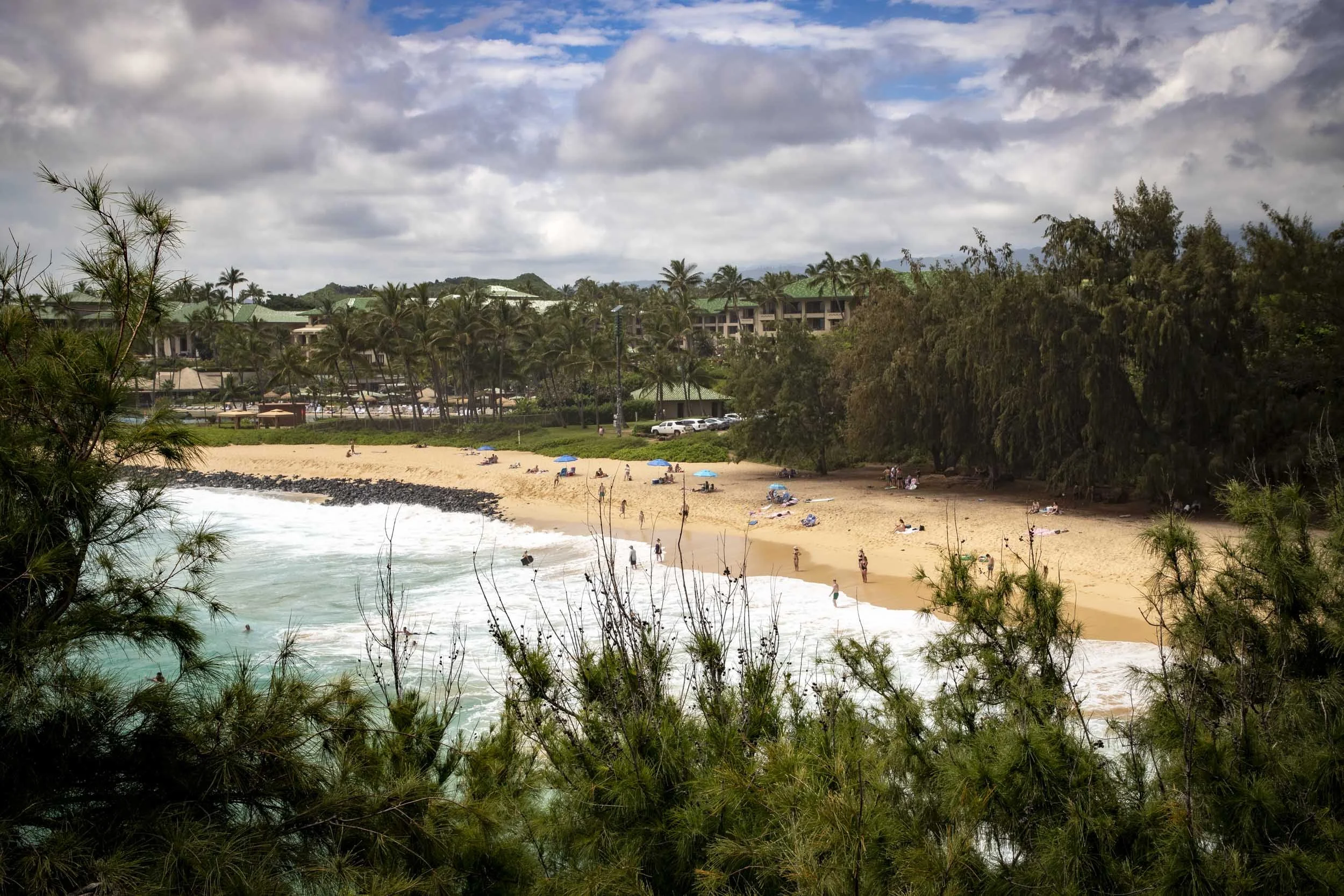 Sandy beach with scattered visitors, turquoise waves, and lush trees under a partly cloudy sky. Shipwreck Beach, Kauai, Hawaii