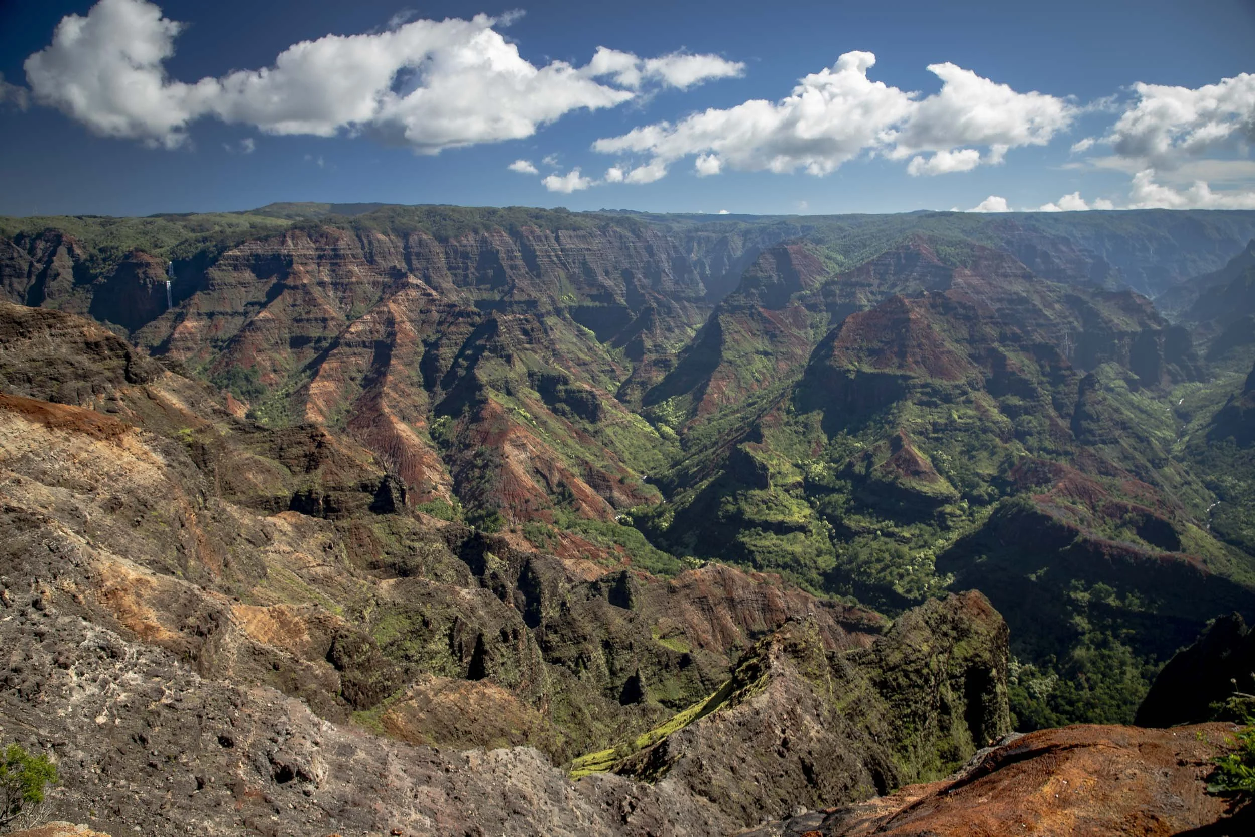 Vast red and green canyon with rugged cliffs and a distant waterfall under a bright blue sky. Waimea Canyon, Kauai, Hawaii.