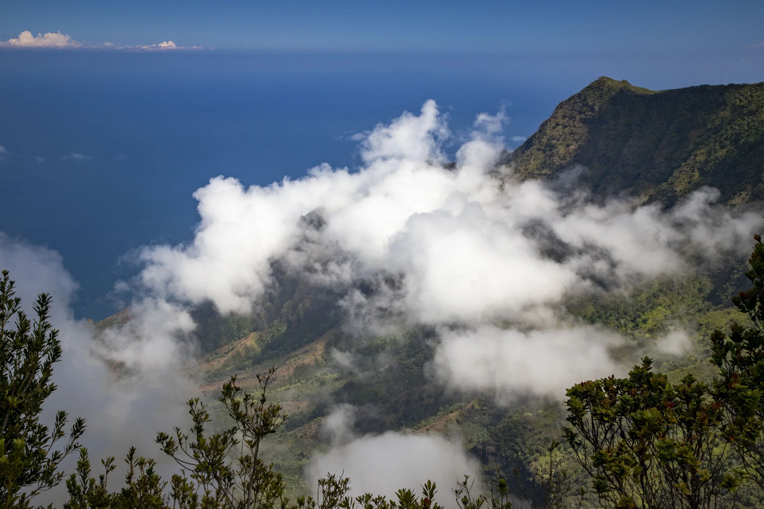 Clouds drift over lush green mountains with the blue ocean beyond, creating a serene tropical view. Location: Waimea Canyon, Hawaii.