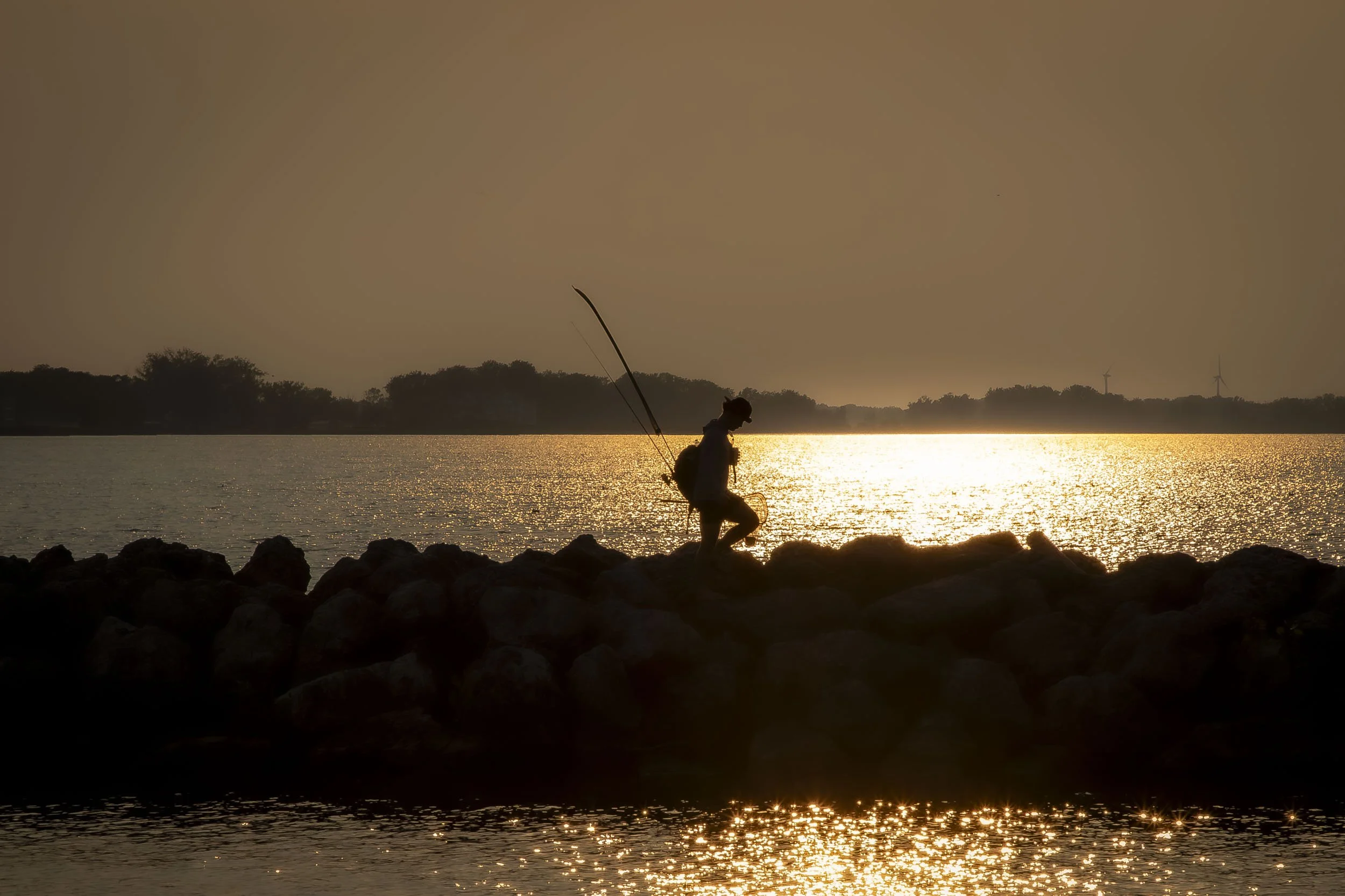 A silhouette of a person fishing by a body of water during sunset, with wind turbines and trees in the background.