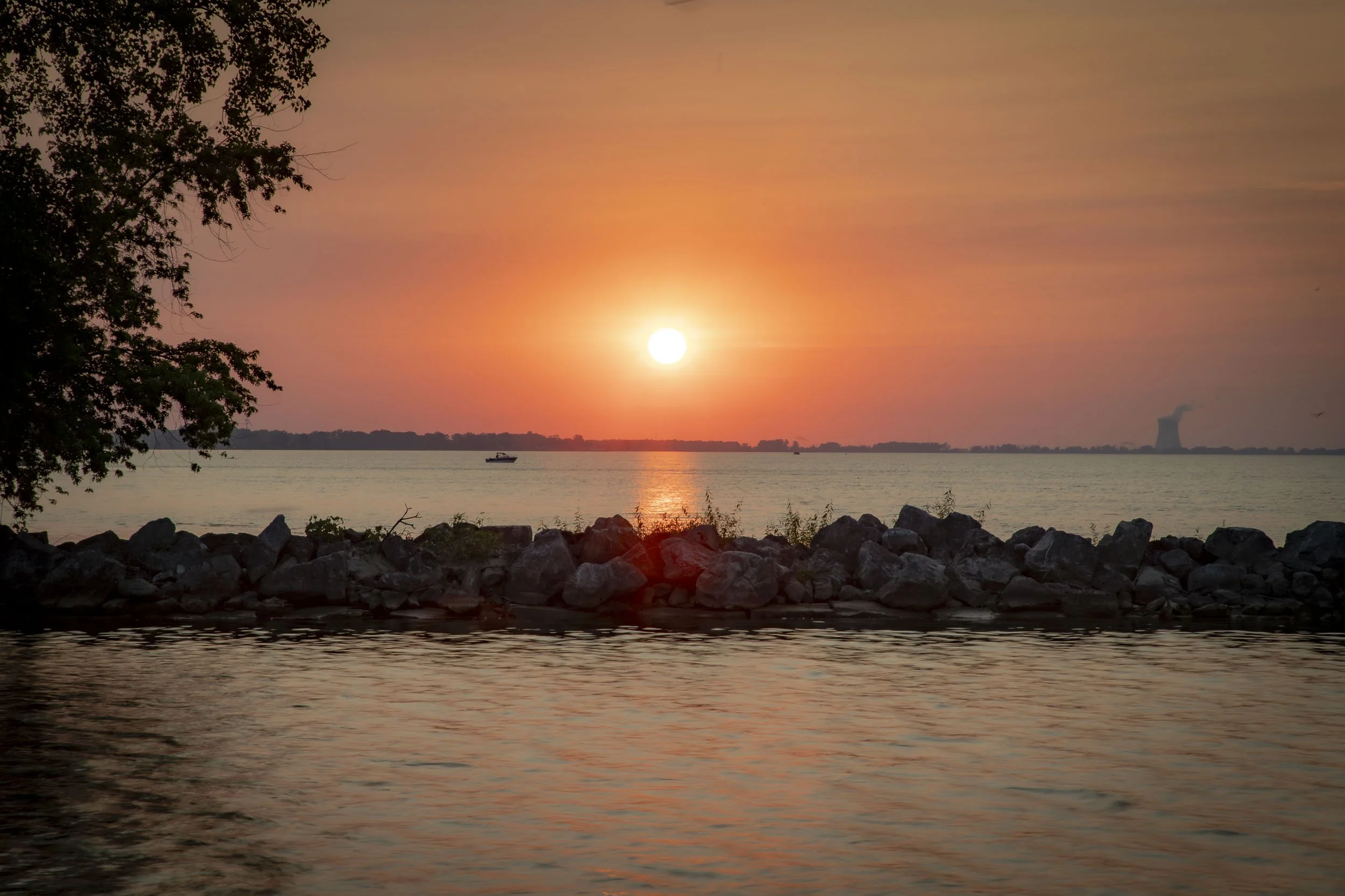 A sunset over a body of water, with a boat in the distance, rocks along the shoreline, and trees silhouetted on the left.