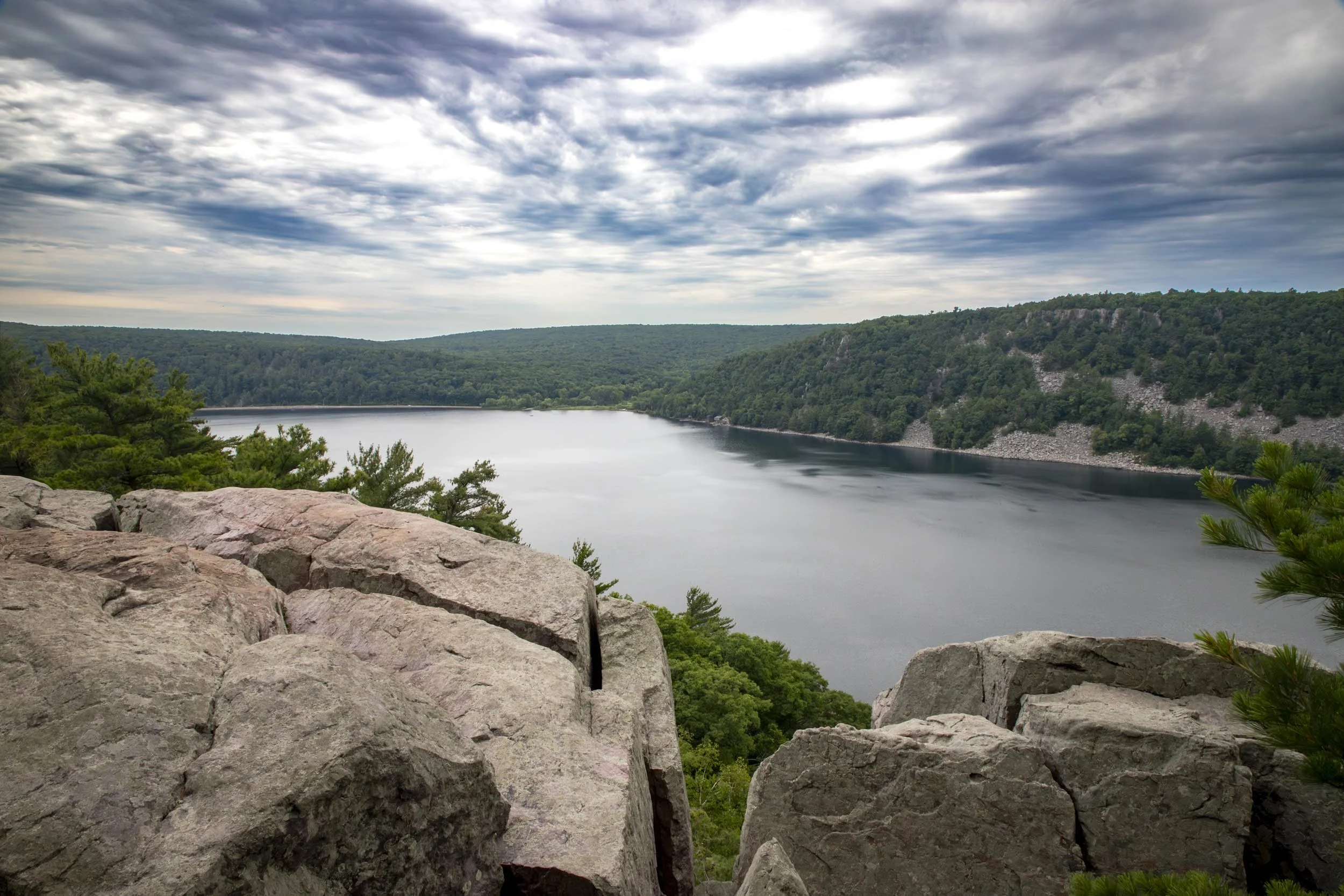 Rocky cliffs overlook a serene lake surrounded by dense green forest under a cloudy sky. Devil's Lake State Park, Wisconsin.