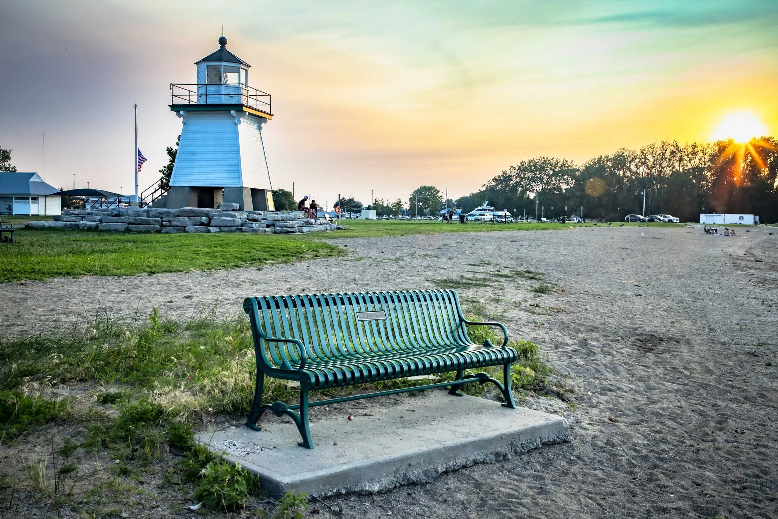 The image depicts a lighthouse at sunset, with a green bench on a sandy beach, and a rocky shoreline.