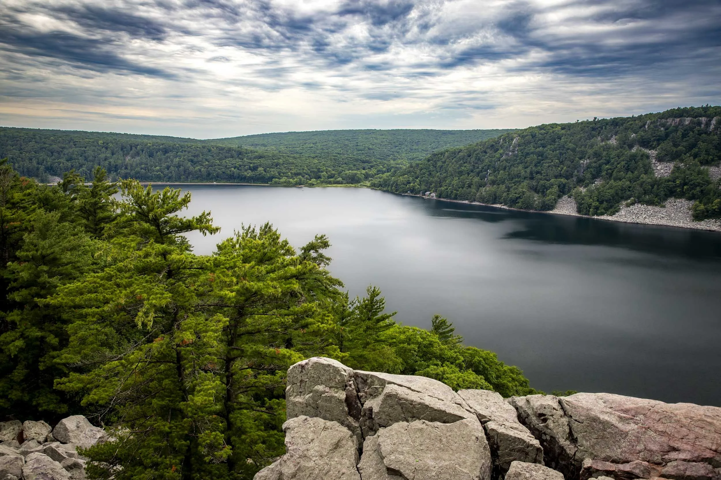 Rocky cliffs overlook a calm lake surrounded by dense green forest under a cloudy sky. Location: Devil's Lake State Park, Wisconsin.
