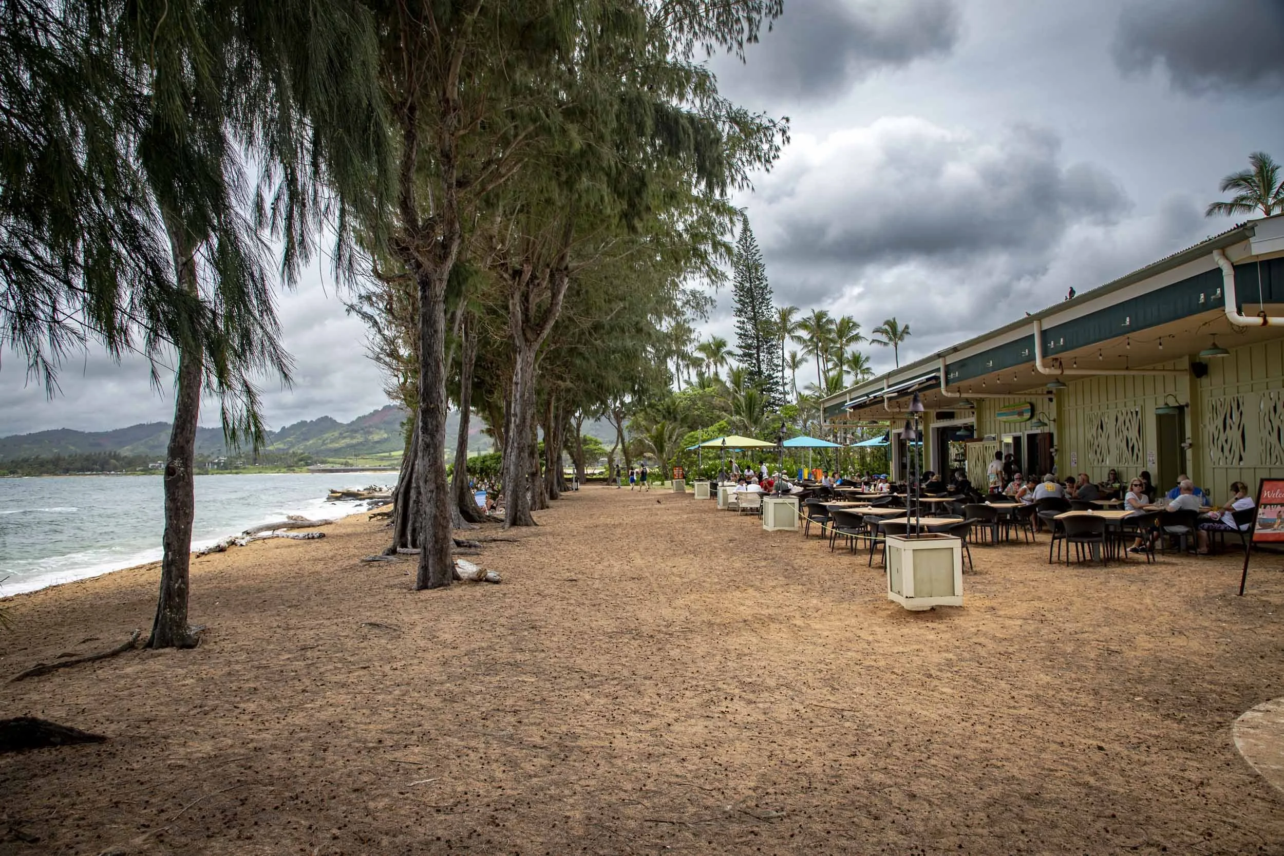 Beachside café with outdoor seating under cloudy skies, lined with tall trees by the ocean. Wailua Beach, Kauai, Hawaii