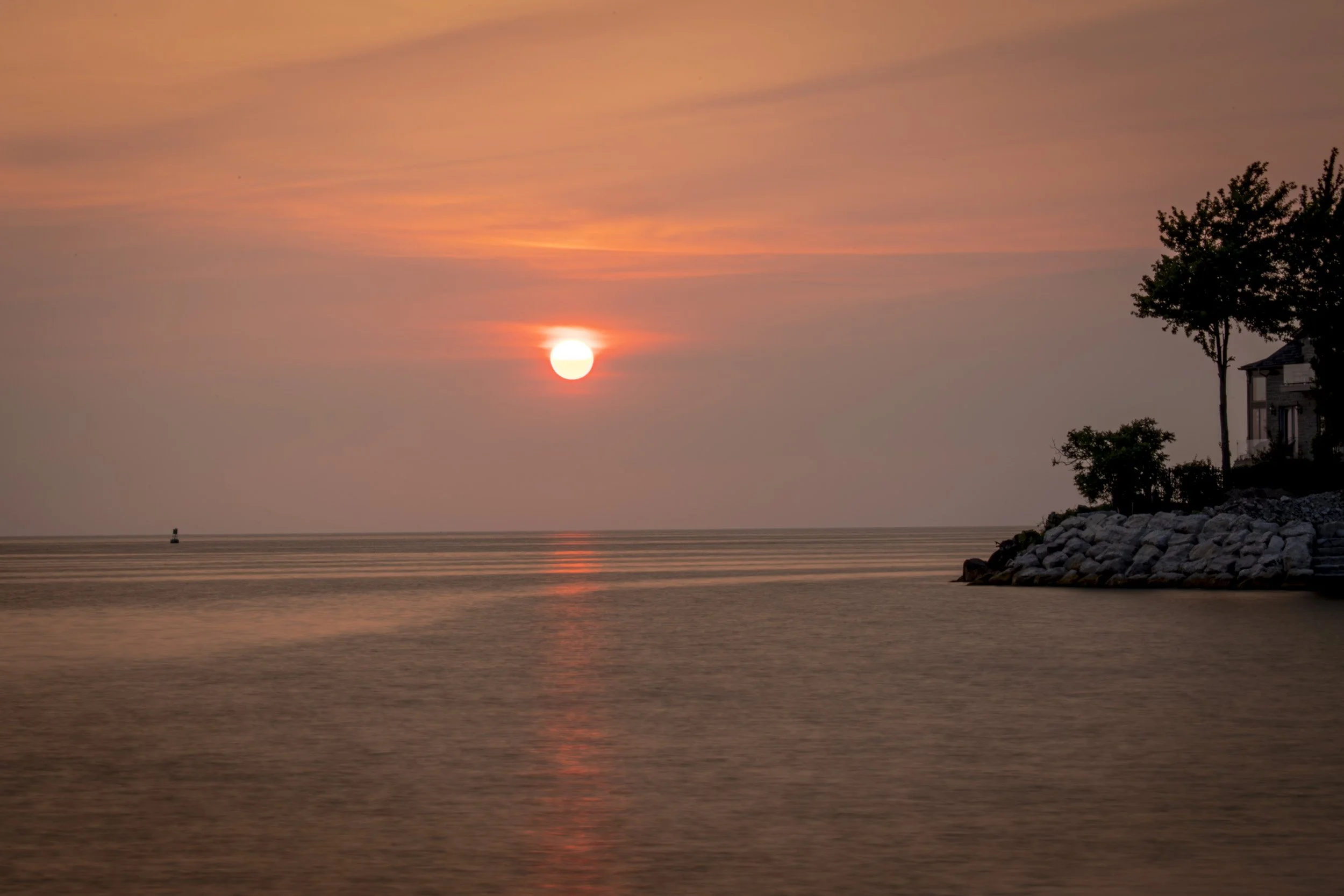 Sunset over a body of water with a small lighthouse in the distance, rocks and trees on the shore on the right side, colorful sky with pink and orange hues.