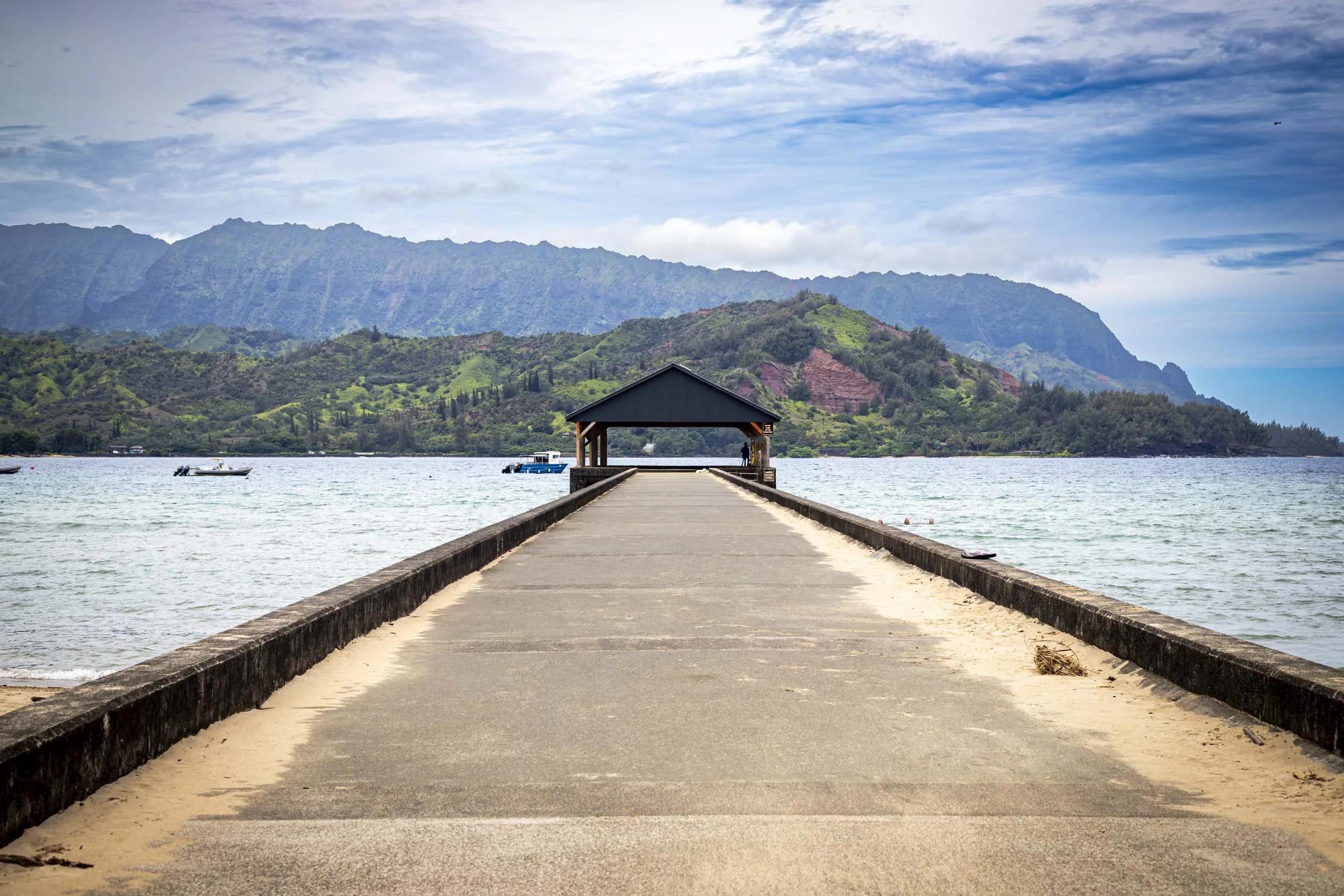A long pier stretches into calm waters with lush green mountains in the background. Hanalei Pier, Kauai, Hawaii