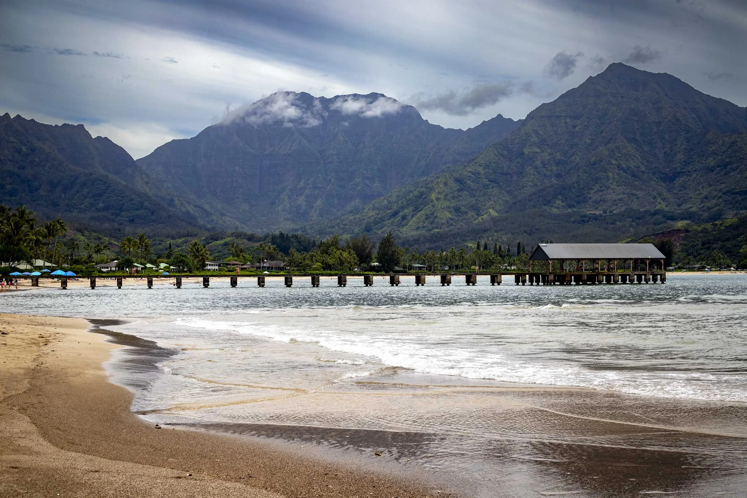 Scenic beach with a long pier, calm waves, and lush mountains under a cloudy sky.