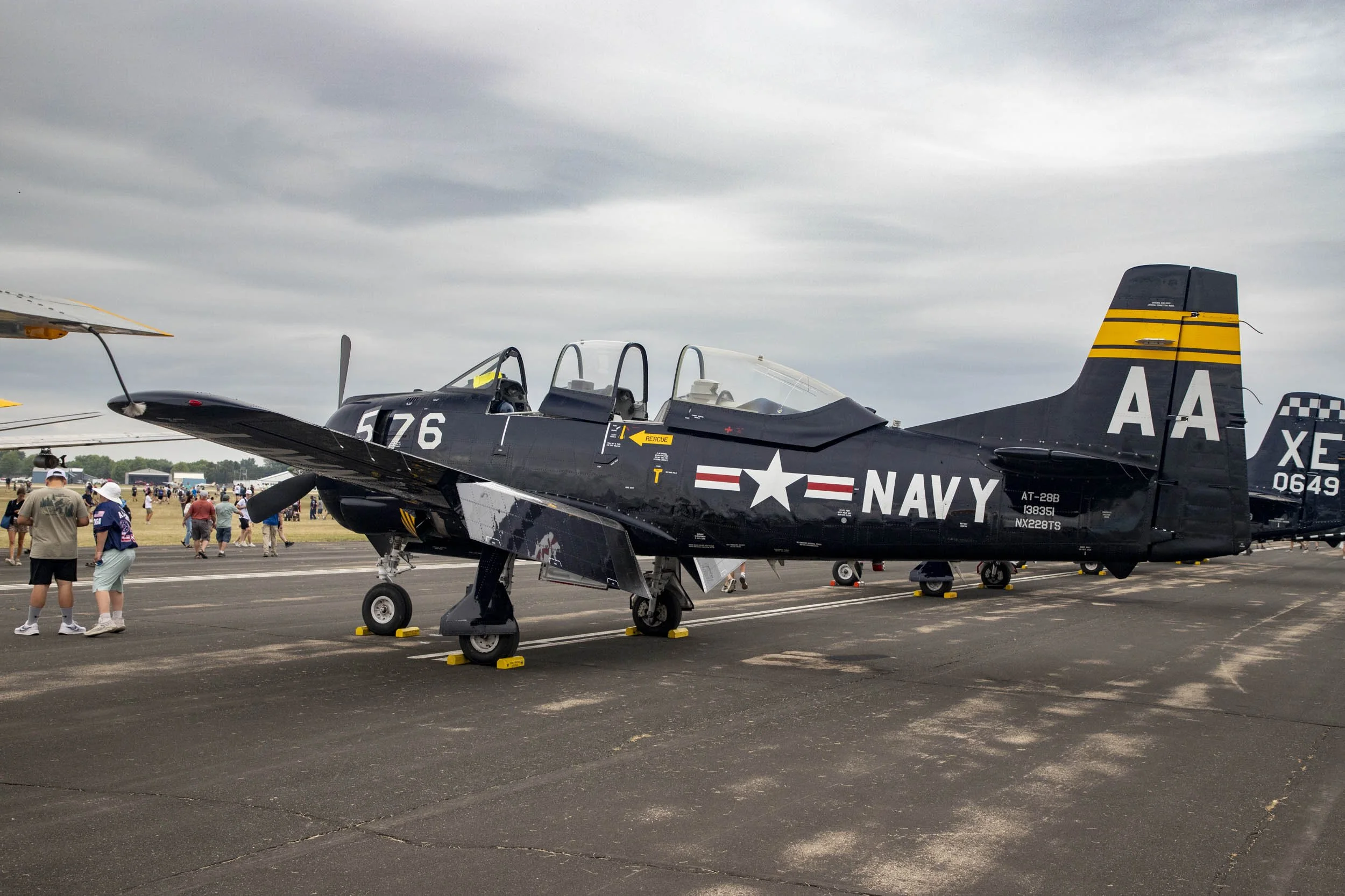 This image features a vintage military trainer aircraft—a North American T-28 Trojan (marked as AT-28B near the tail)—parked on an asphalt tarmac during an airshow event under an overcast sky.