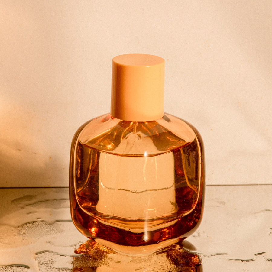 A pink-tinted perfume bottle with a pink cap on a reflective surface against a beige background.