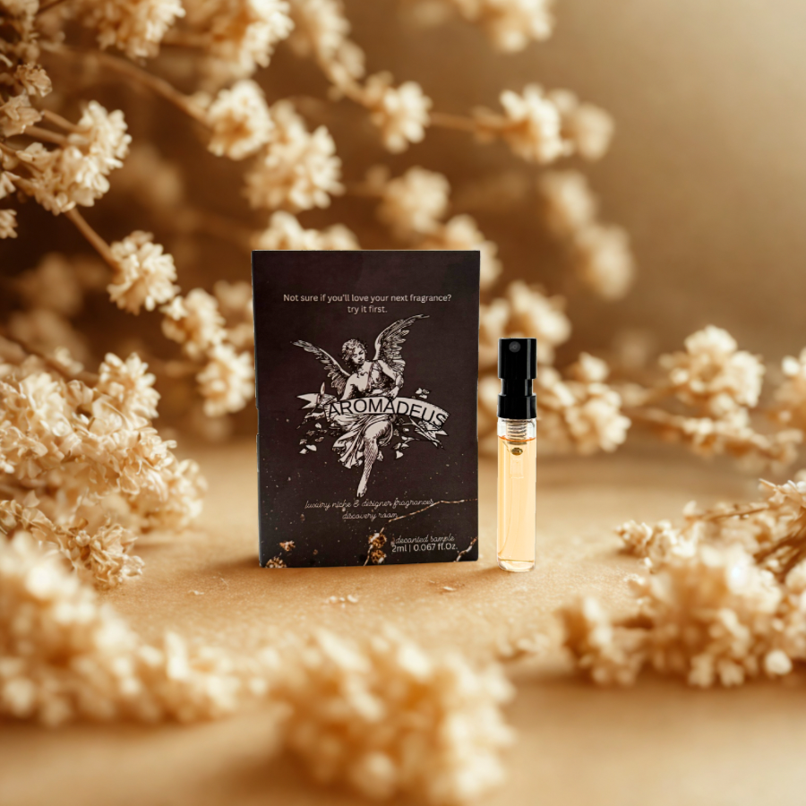 A small perfume sample spray vial with a black cap, placed beside a black packet labeled "AROMADEUS" with a vintage angel illustration, surrounded by dried white flowers on a beige background.