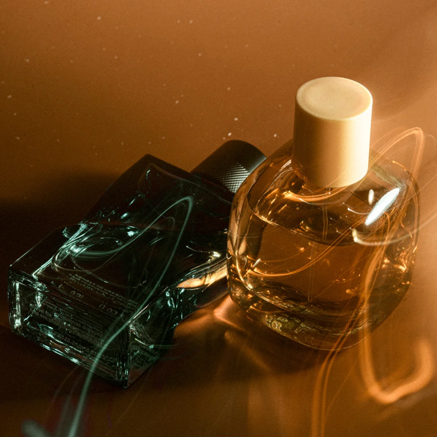 Two perfume bottles, one black and rectangular, the other round and clear, placed on a reflective surface with warm lighting creating reflections and light streaks.