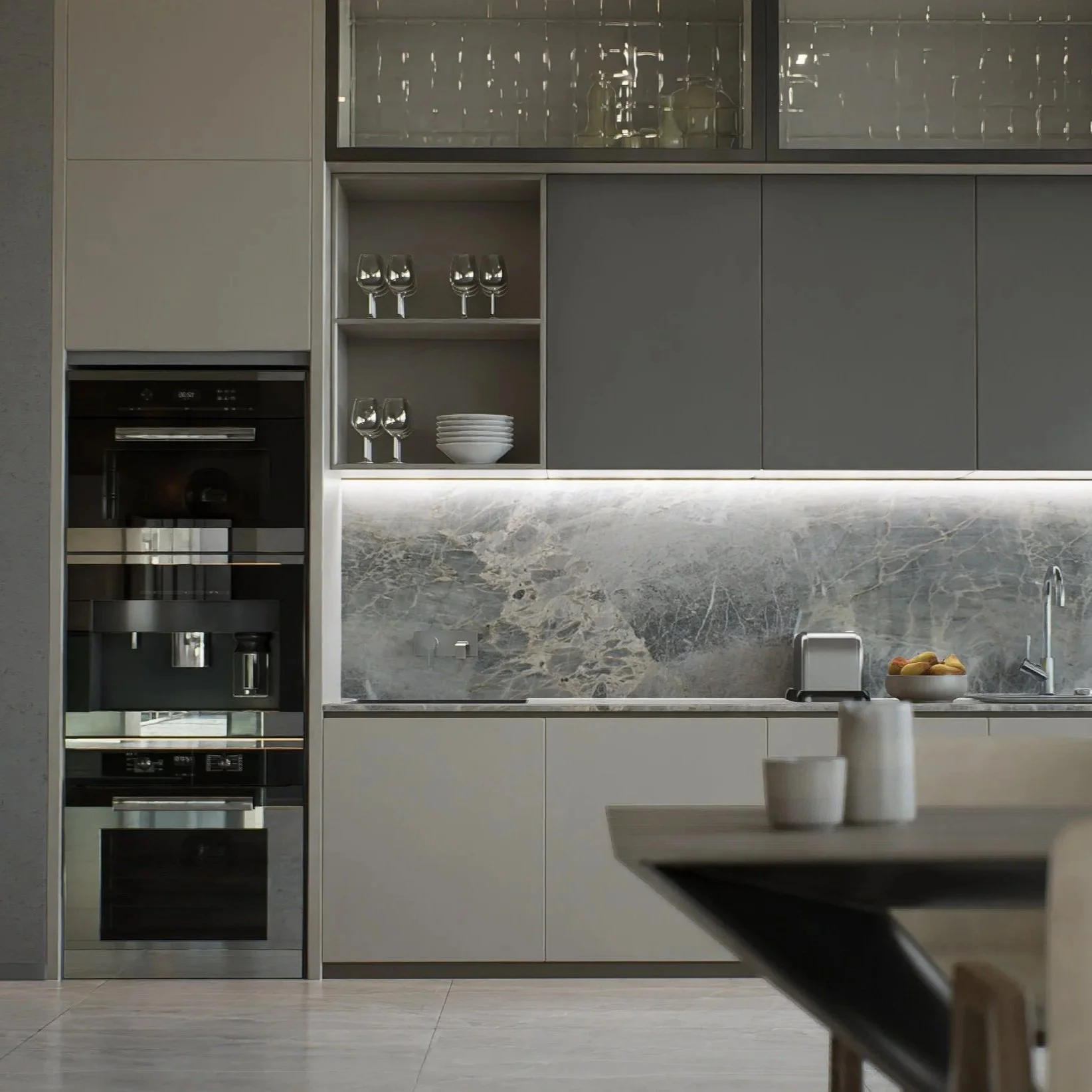 Modern kitchen with gray cabinets, a marble backsplash, and built-in appliances. Glassware and bowls are arranged on open shelves, and a bowl of fruit is on the countertop. A table with white vases and cups is partially visible in the foreground.