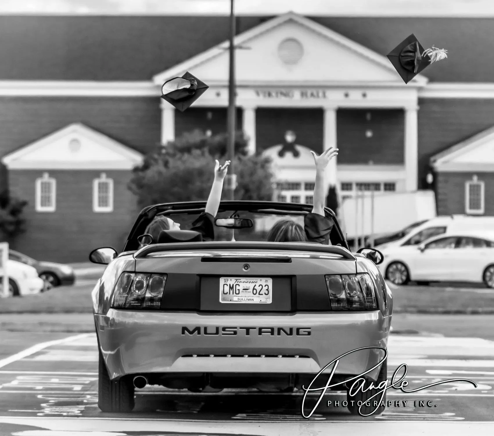 Two graduates in caps and gowns celebrating in a convertible Mustang car, tossing graduation caps into the air in a parking lot with a school building in the background.
