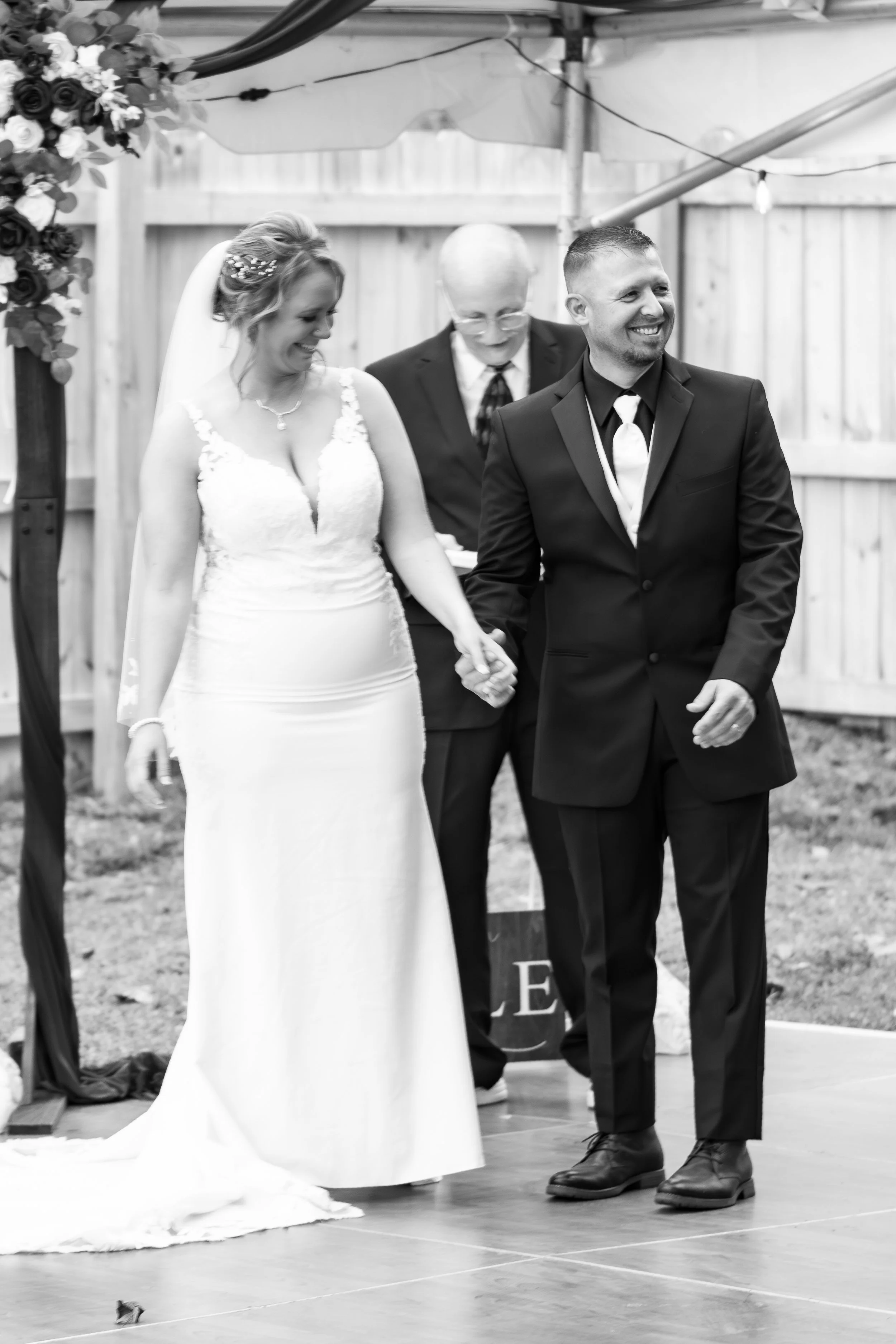 A bride and groom holding hands, smiling during their wedding ceremony outdoors. An officiant is standing behind them, in front of a wooden fence and decorated arch.