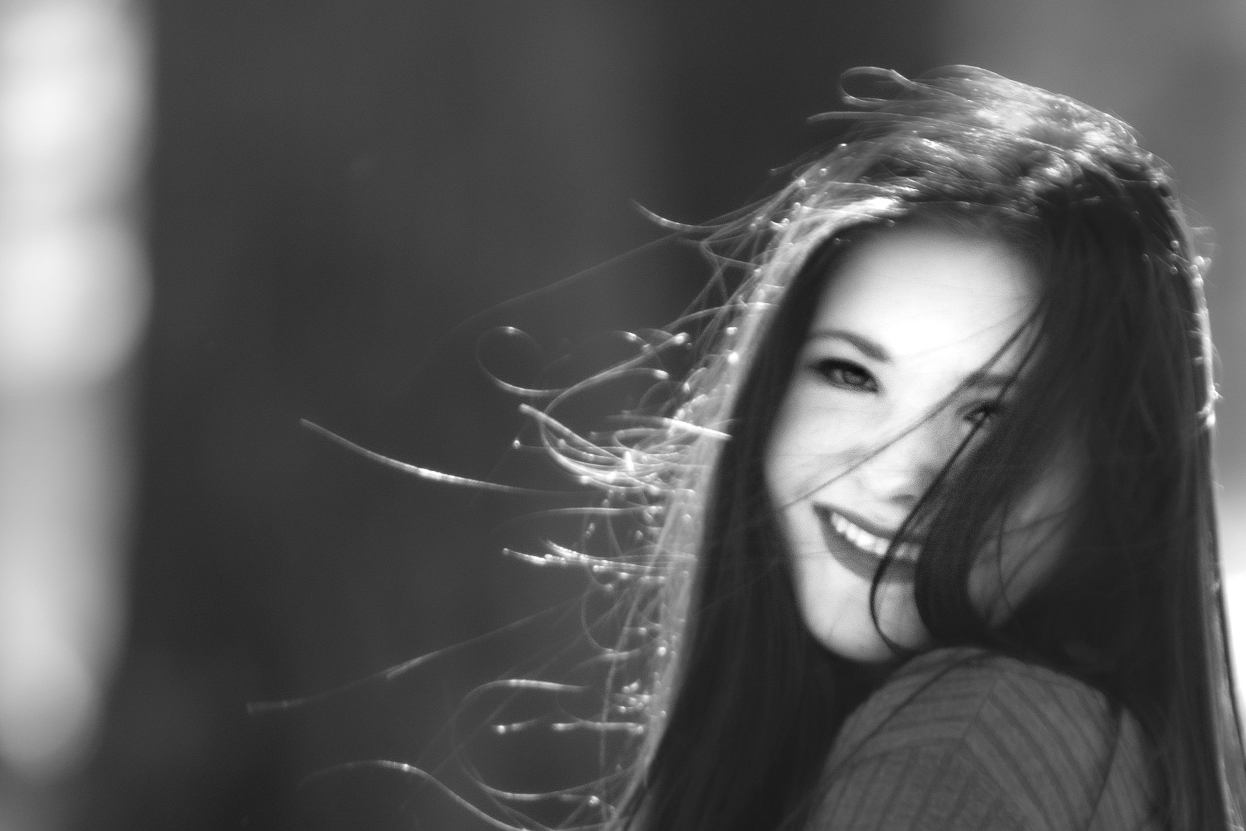 A black-and-white photo of a young woman with long hair, smiling and looking over her shoulder, with her hair blowing in the wind.