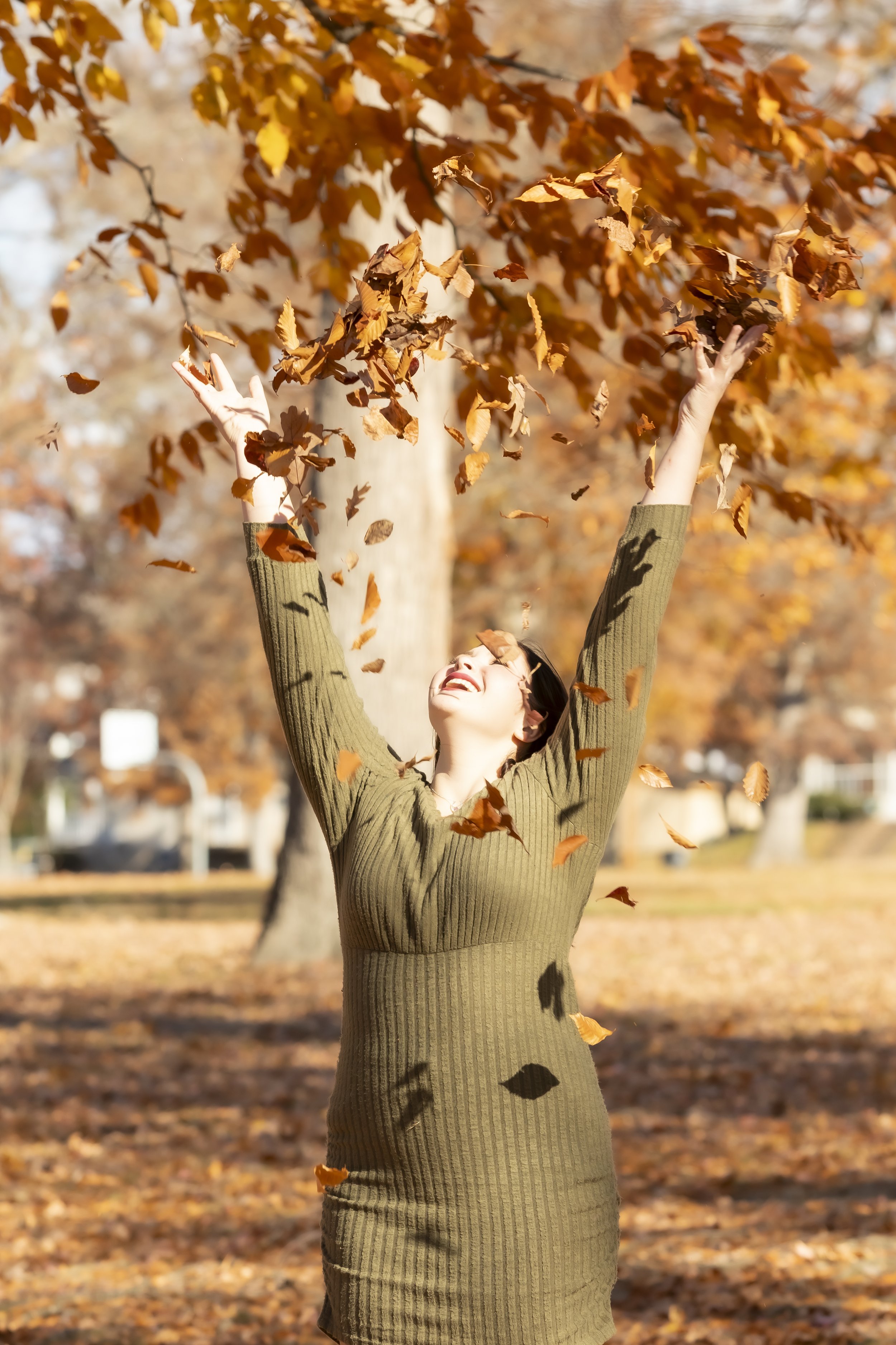 A woman smiling and playing with falling leaves in a park during autumn.