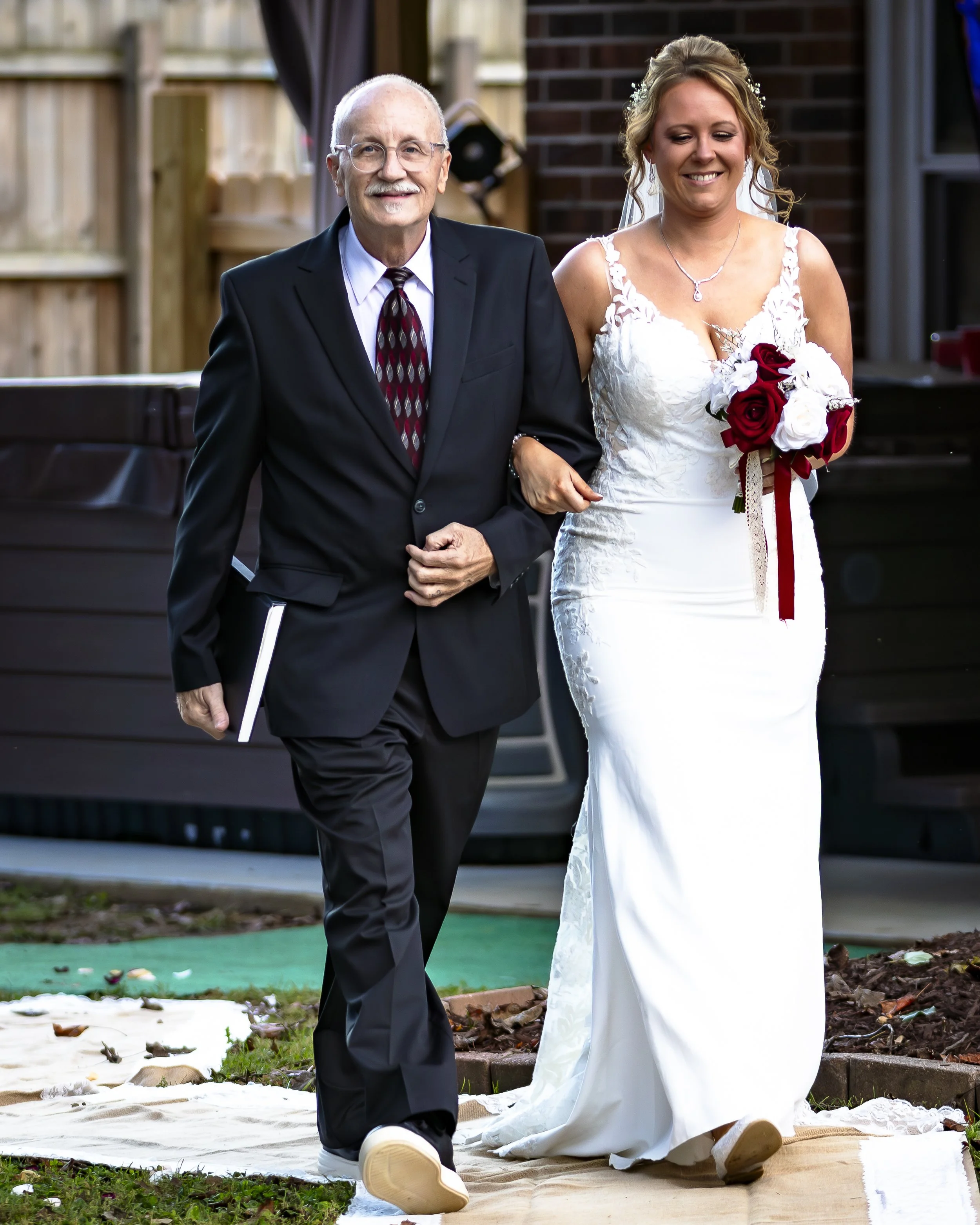A bride in a white wedding dress holding a bouquet of red and white roses walks arm-in-arm with an older man in a black suit, during a wedding ceremony outdoors.