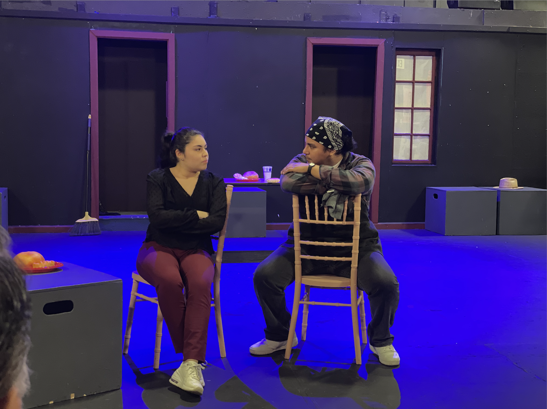 A man sitting on a chair backwards, looking at the girl he loves who is pissed at him. She is sitting with her arms crossed looking at the man.