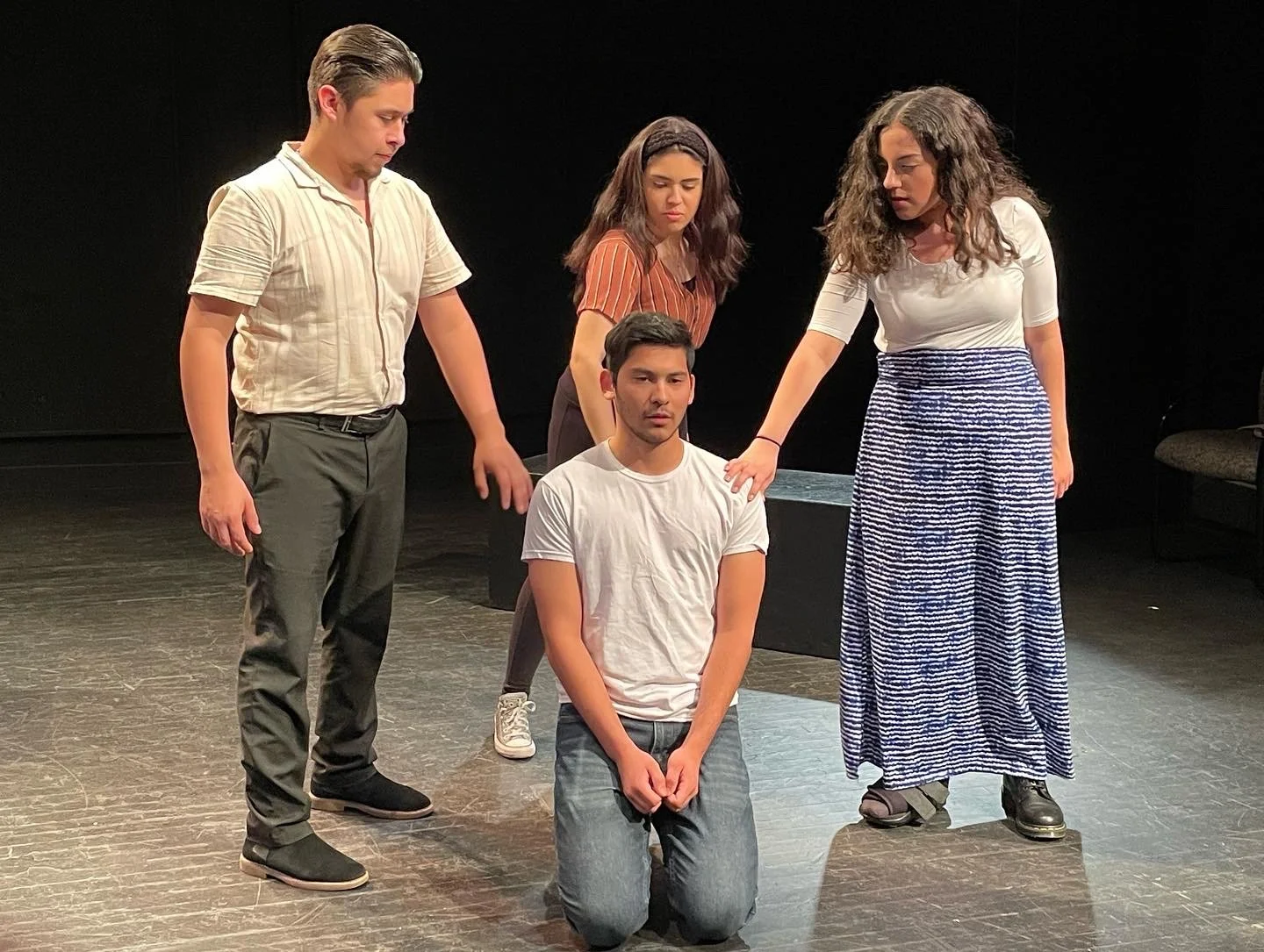A man kneeling on the ground surrounded by his friends, a man and two women, as they lay hands on his shoulders