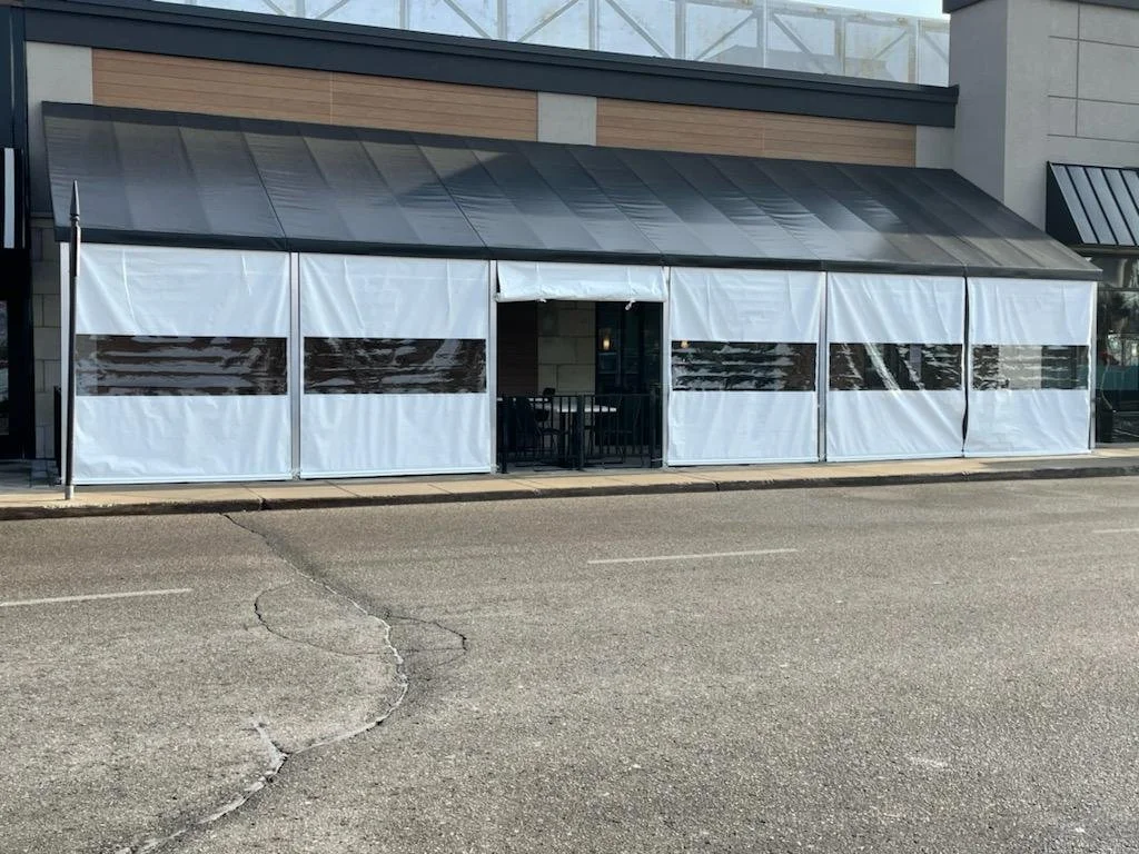 Outdoor seating area with white tent walls at a restaurant or cafe, featuring clear plastic windows, located on a street.