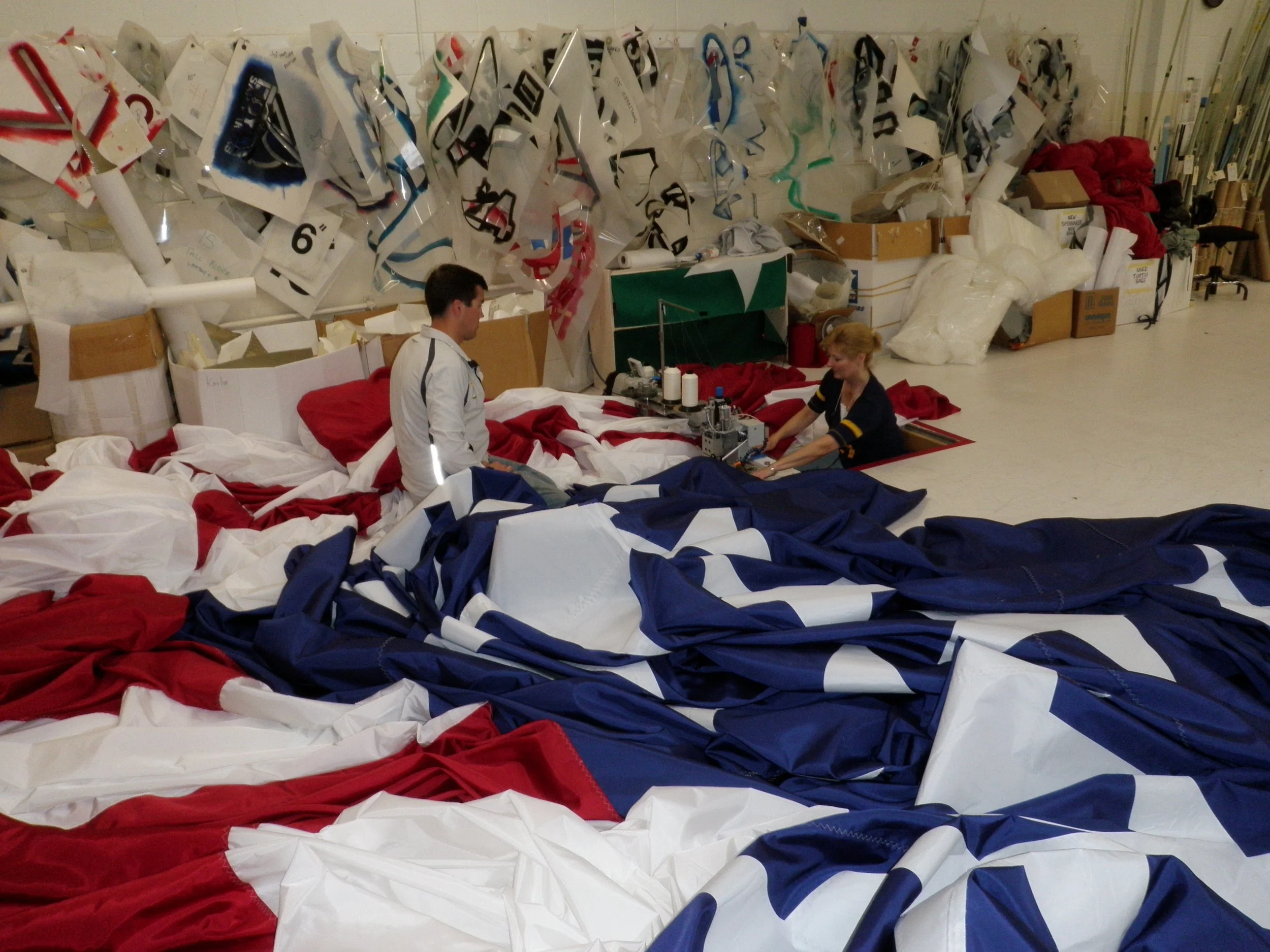 Two people working on sewing machine fabric in a workshop with flags and other materials around them.