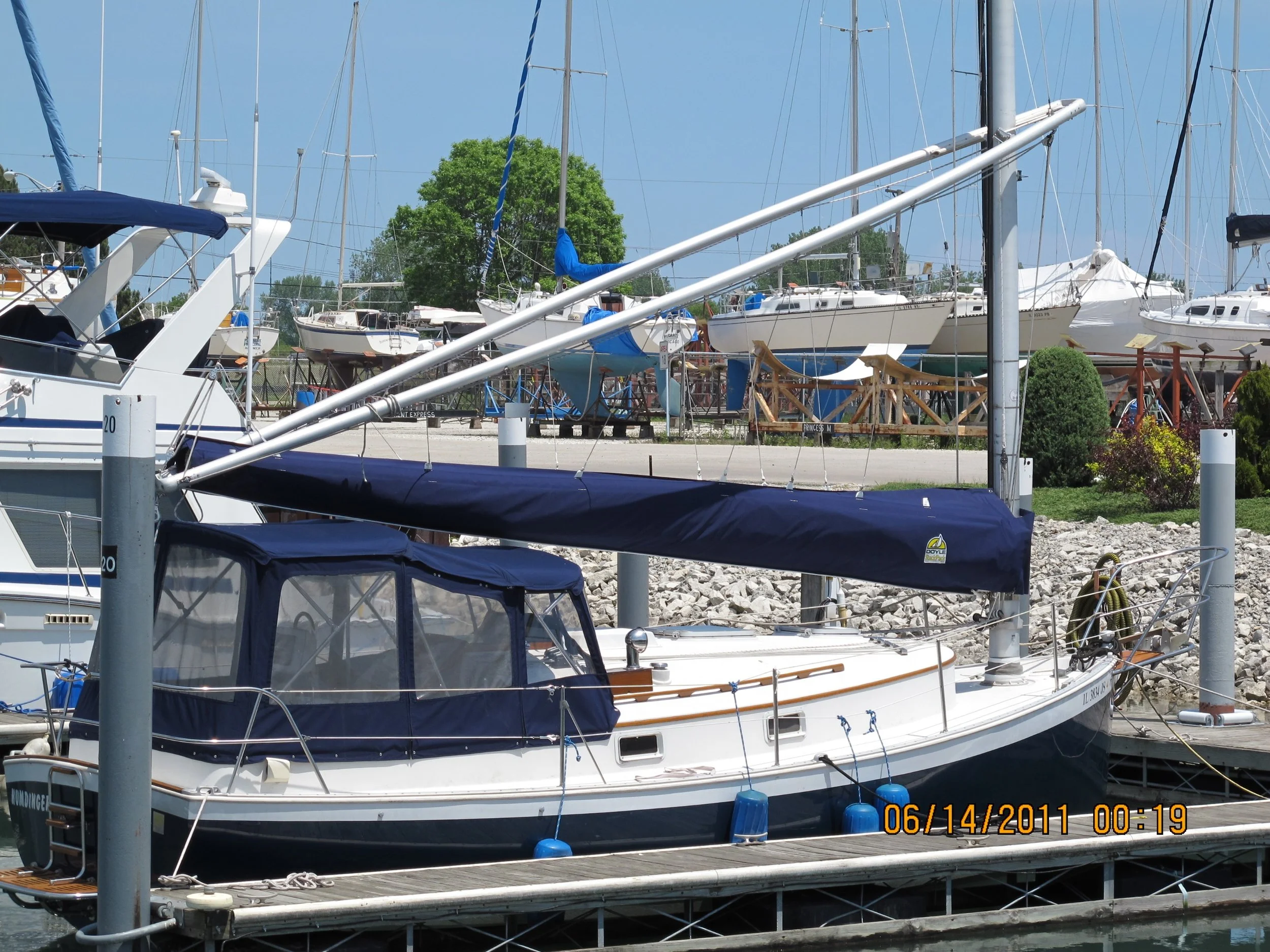 A sailboat docked in a marina with a blue cover over the cockpit area. Other sailboats and powerboats are visible in the background, with some boats on land for maintenance. The date stamp on the image reads June 14, 2011.