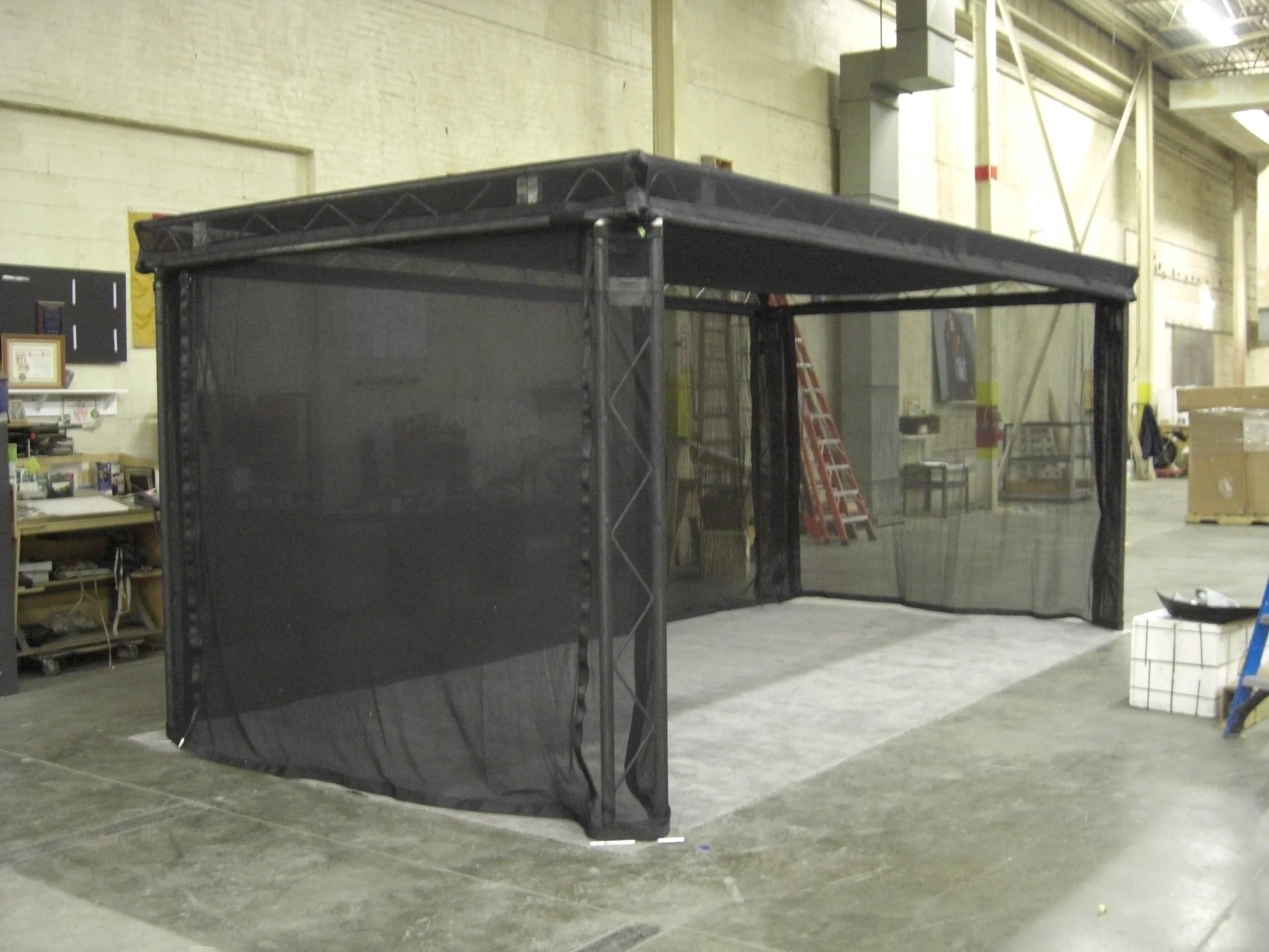 Black fabric structure resembling a booth or enclosure inside an industrial warehouse with visible equipment and tools.