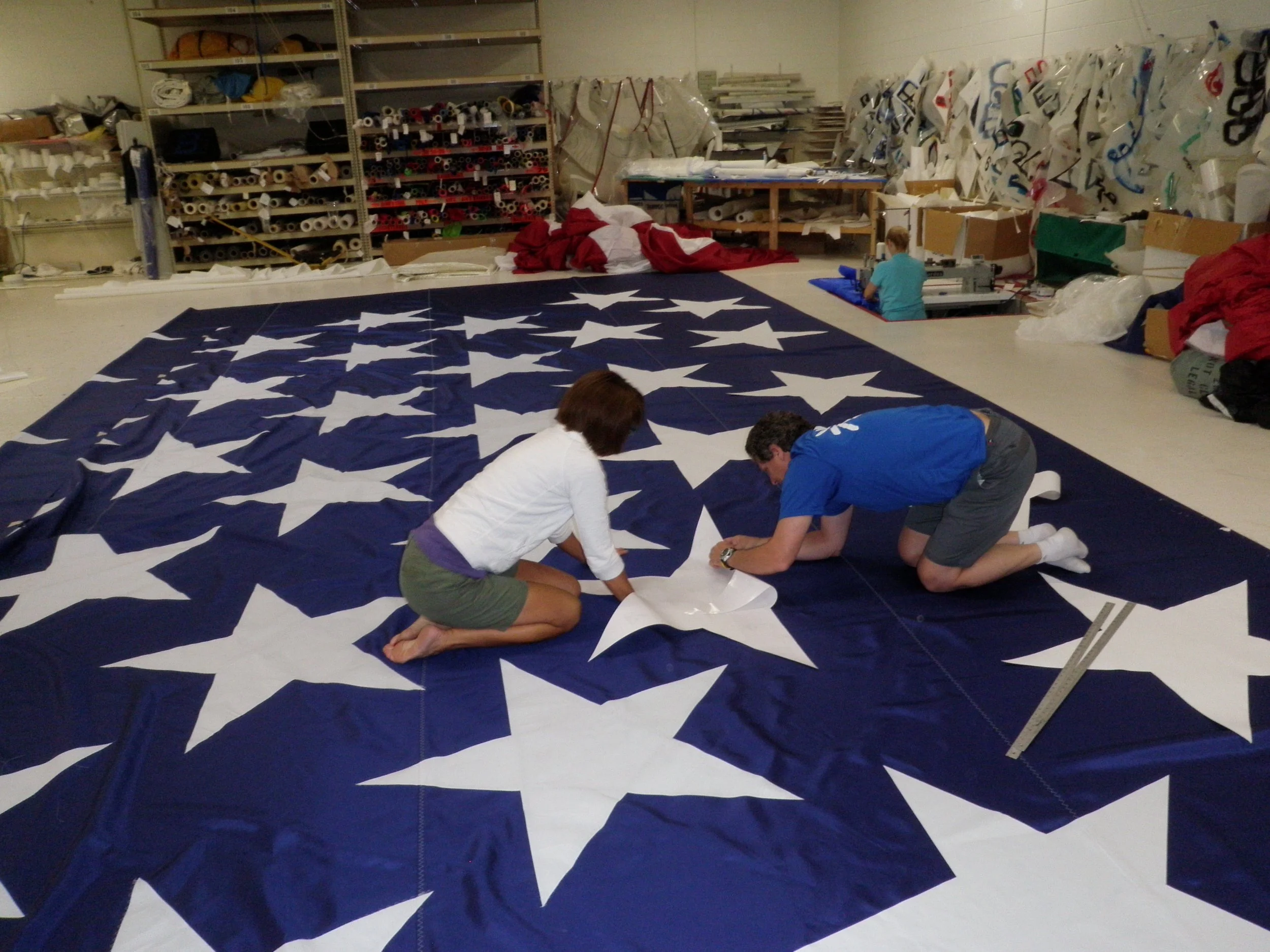 Two people working on a large fabric with white stars on a blue background, similar to a section of the American flag, in a workshop setting with shelves and materials in the background.