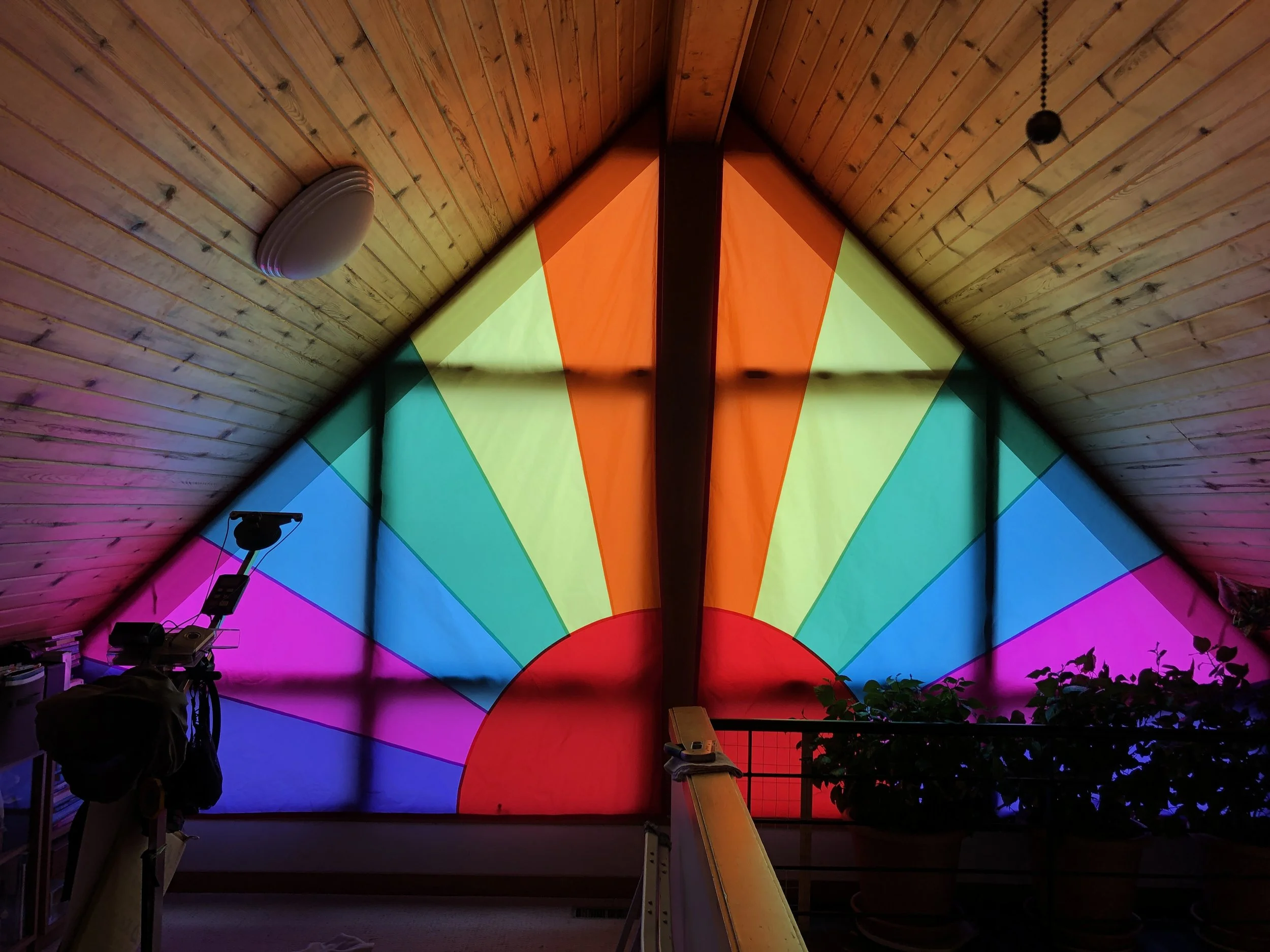 Colorful stained glass window in a loft with rainbow sunburst design, indoor plants, and wooden ceiling.