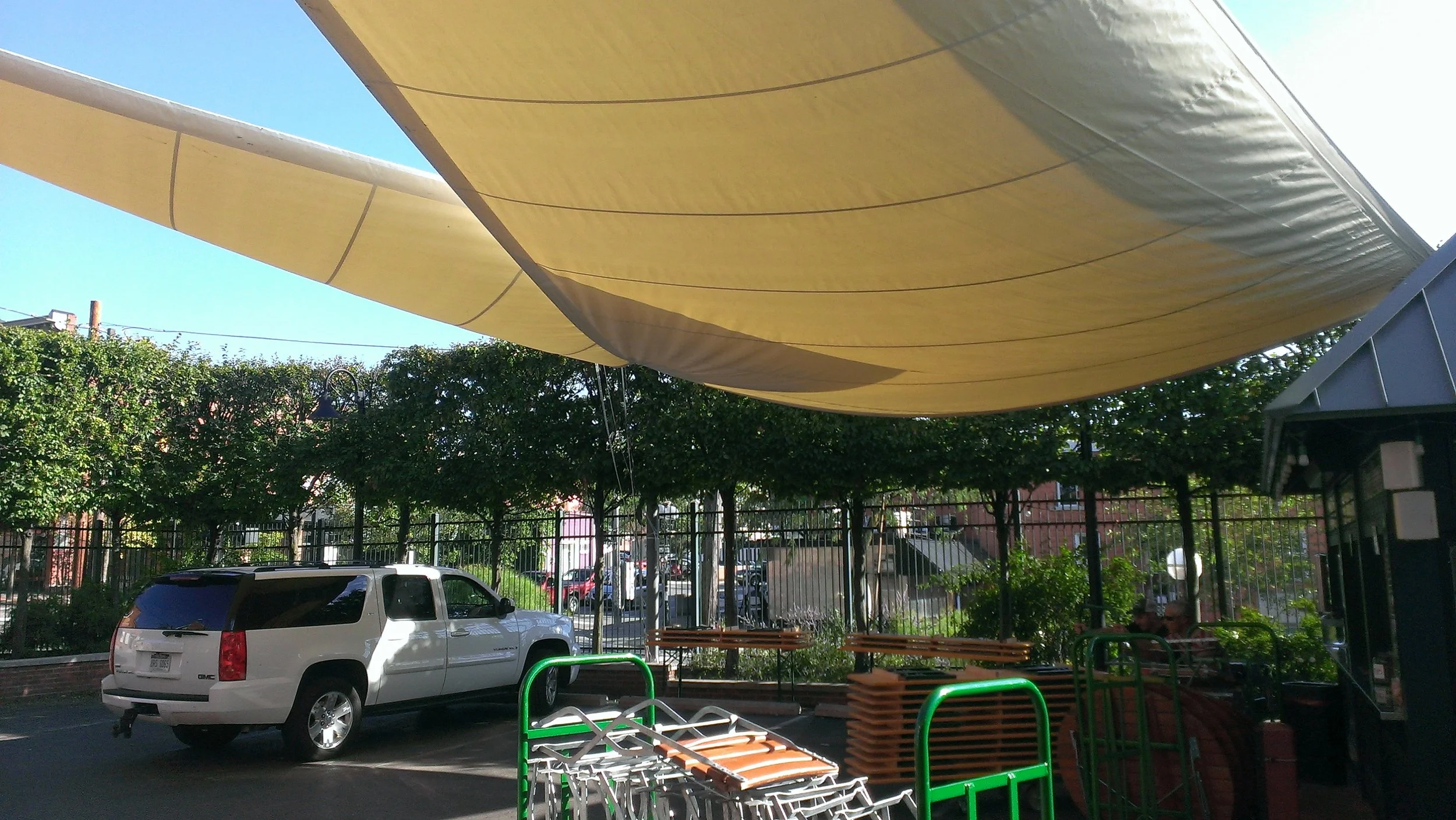 Large white SUV parked under a sunshade structure next to stacked outdoor chairs and trees in a courtyard setting.
