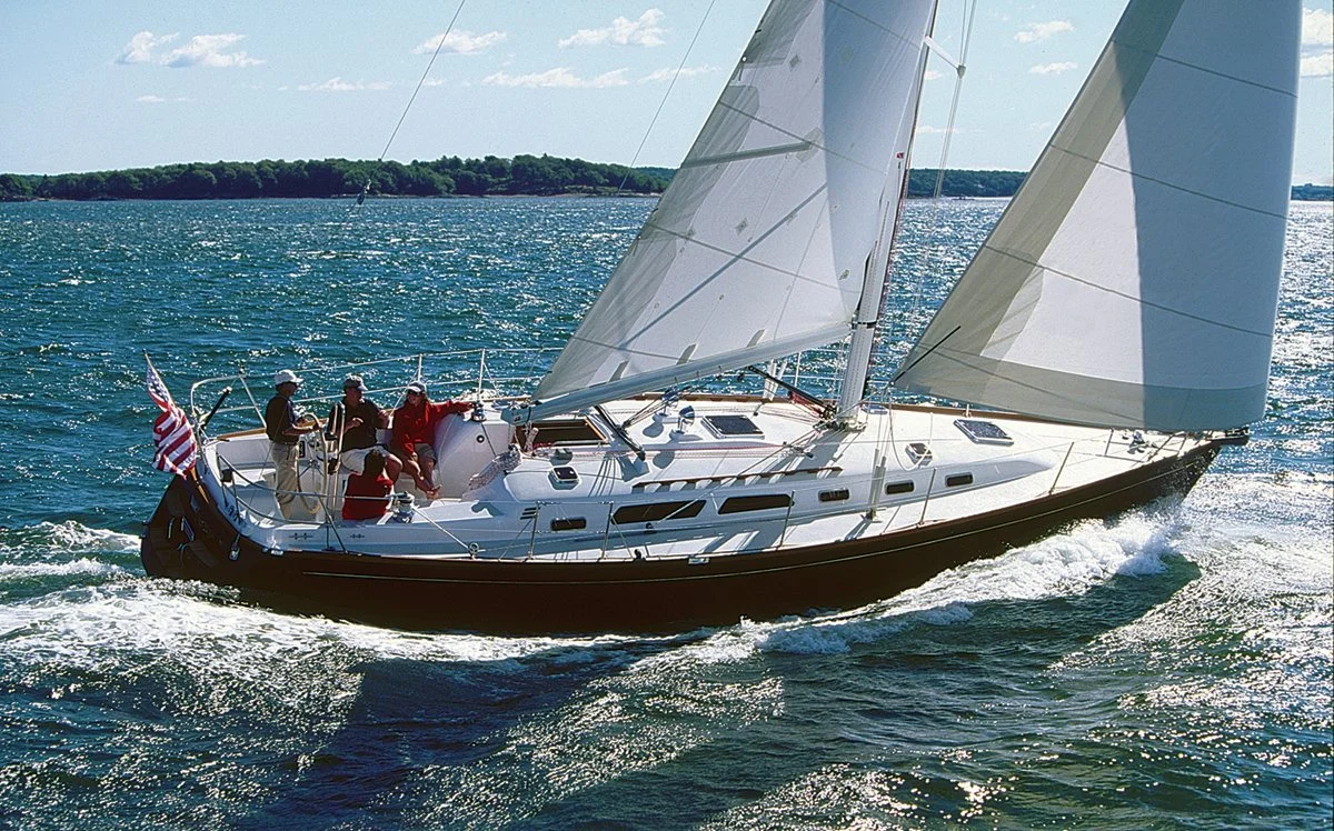 Sailboat with several people on board sailing on a body of water with visible waves and land in the background. The boat is flying an American flag.