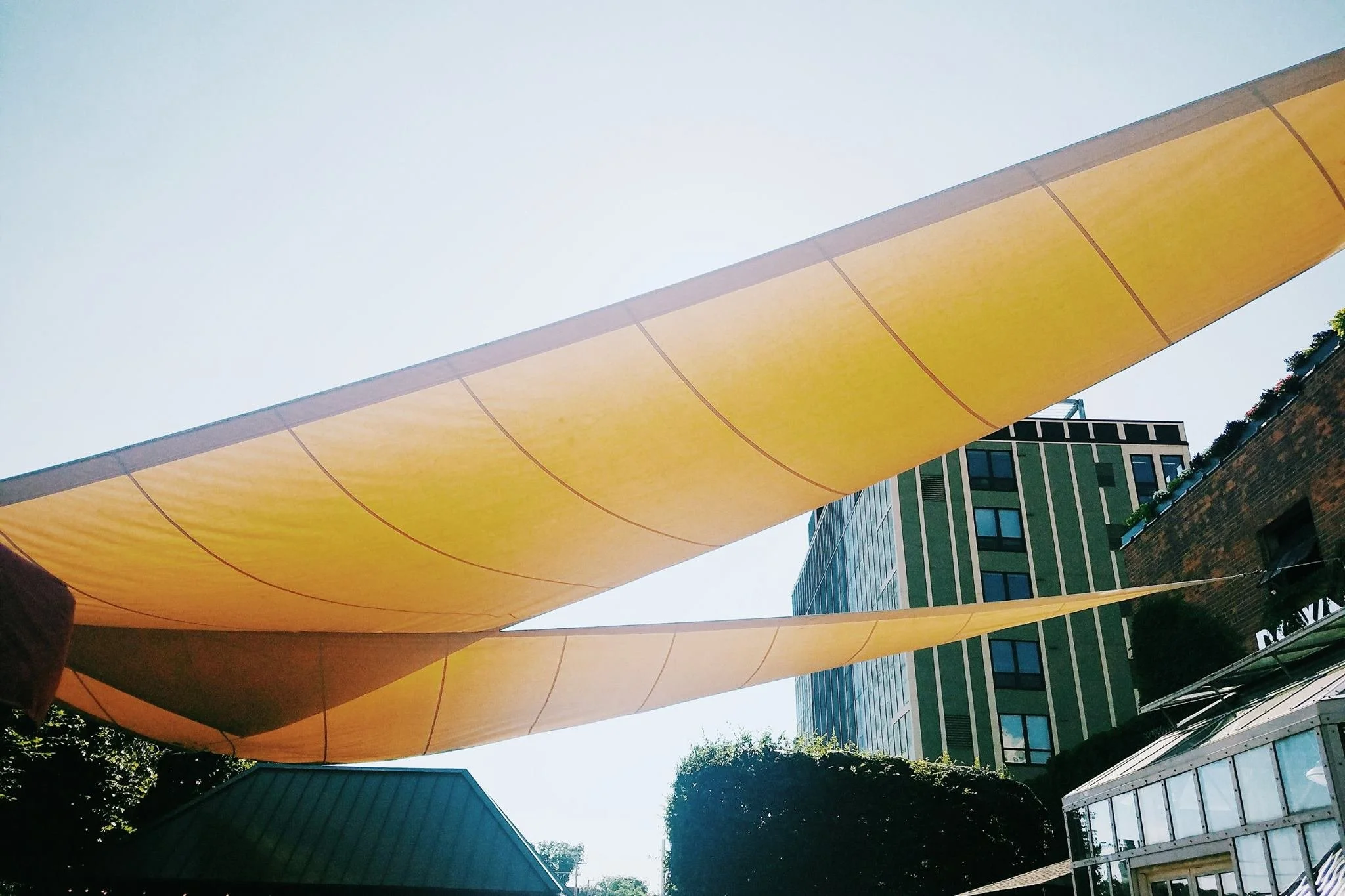 Yellow sunshade sails stretched over an outdoor area with buildings in the background.