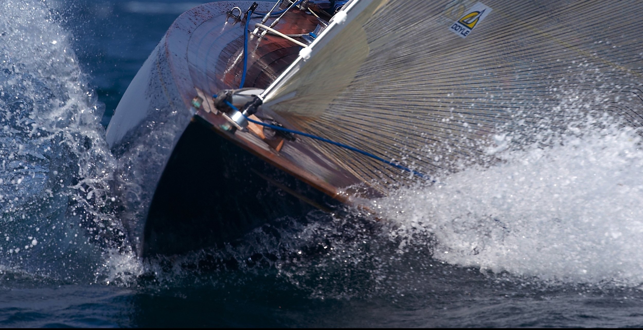 Close-up of a sailboat cutting through water, with splashes around the bow and a visible sail on the right, labeled "doyle."