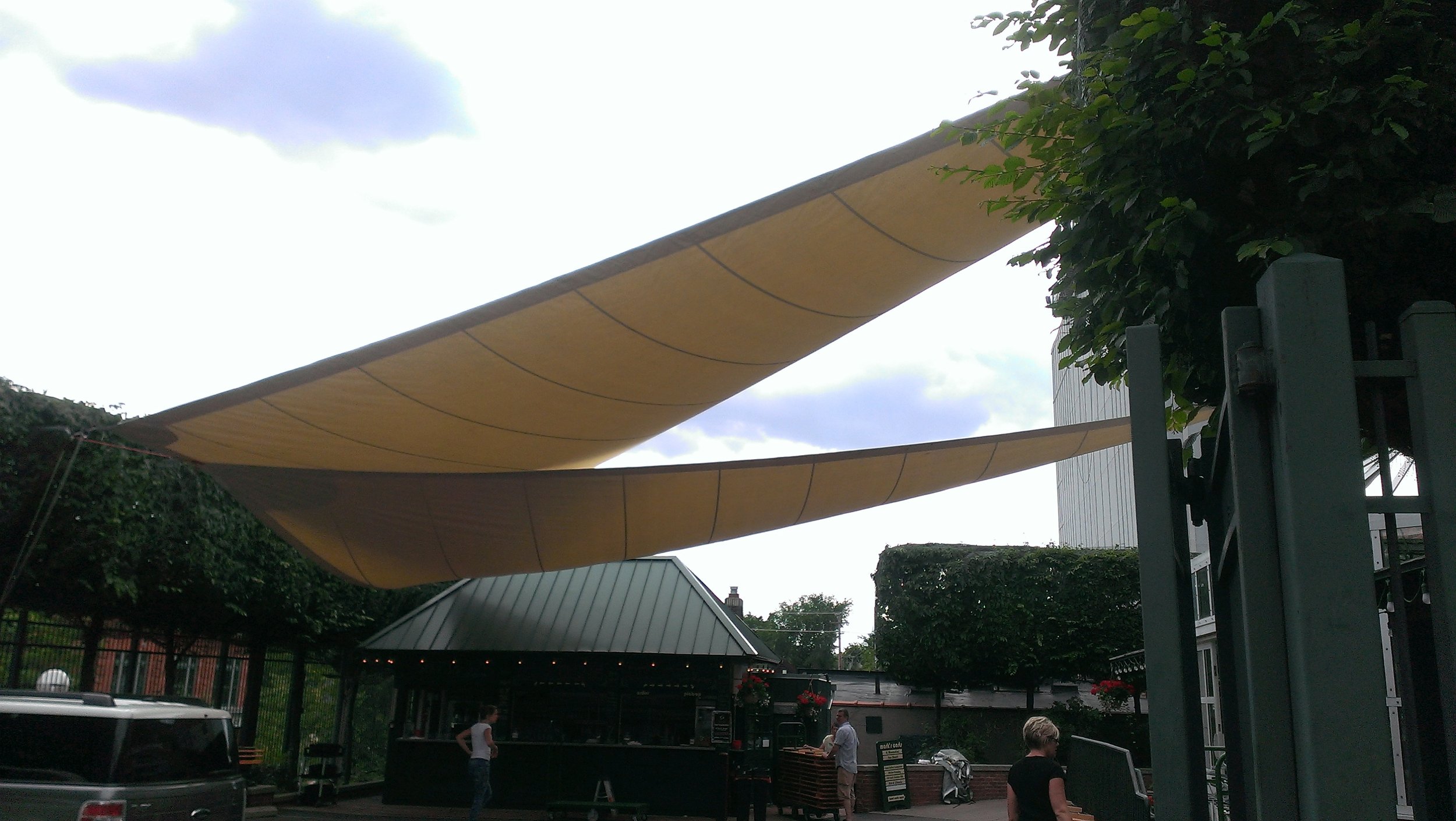 Outdoor patio with yellow sail shade, greenery, people, car, and kiosk.
