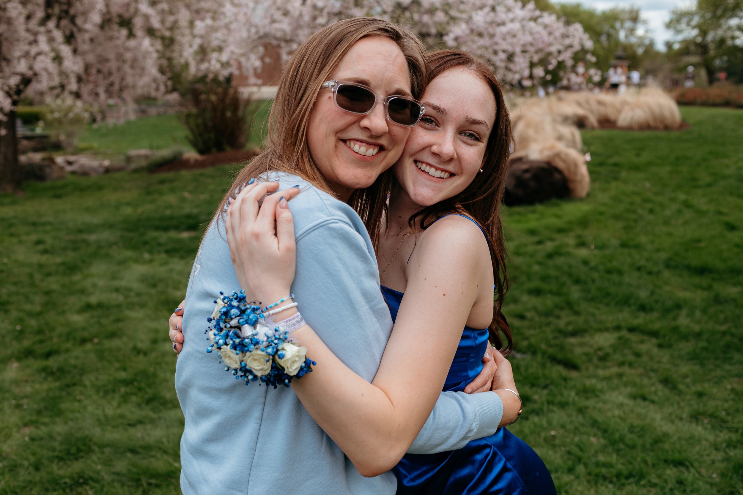 Two people hugging and smiling outdoors, one wearing a blue dress and the other wearing sunglasses and a light blue top, with a corsage on their wrist, surrounded by grass and cherry blossom trees.
