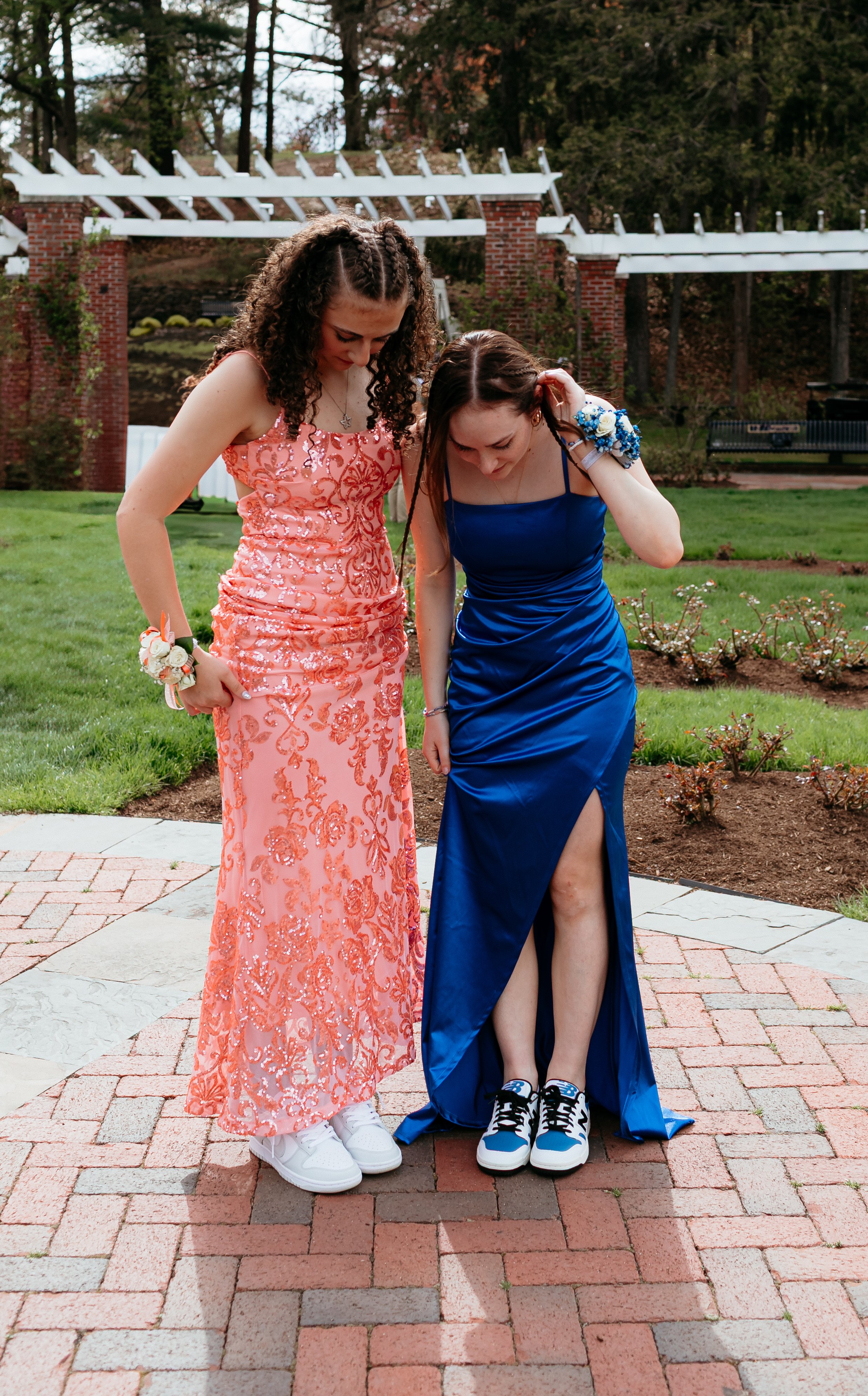 Two people in formal dresses and sneakers stand on a brick walkway, one in a pink sequined gown and the other in a blue satin gown, both wearing corsages.